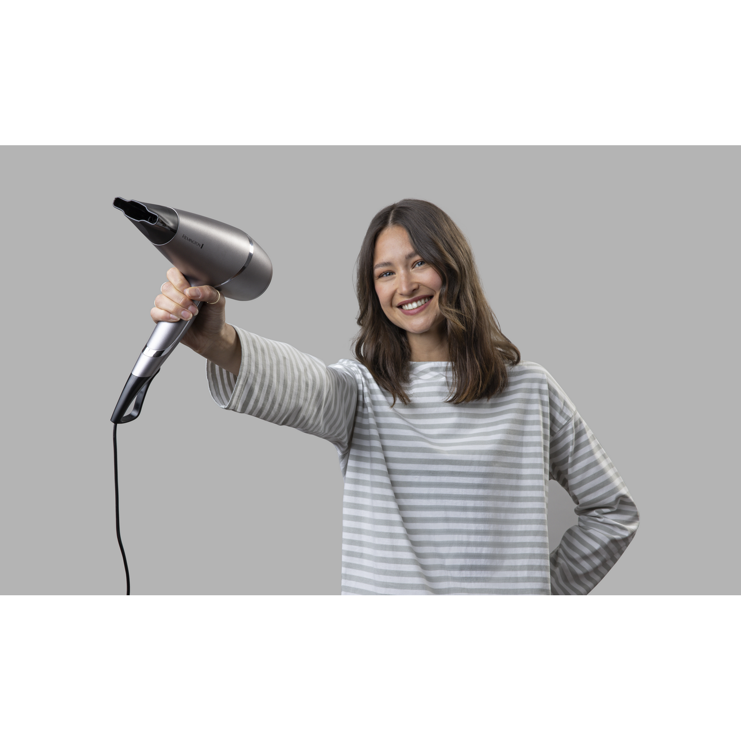 Mujer sosteniendo un secador de pelo plateado, sonriendo, sobre un fondo gris. Lleva una camisa a rayas.