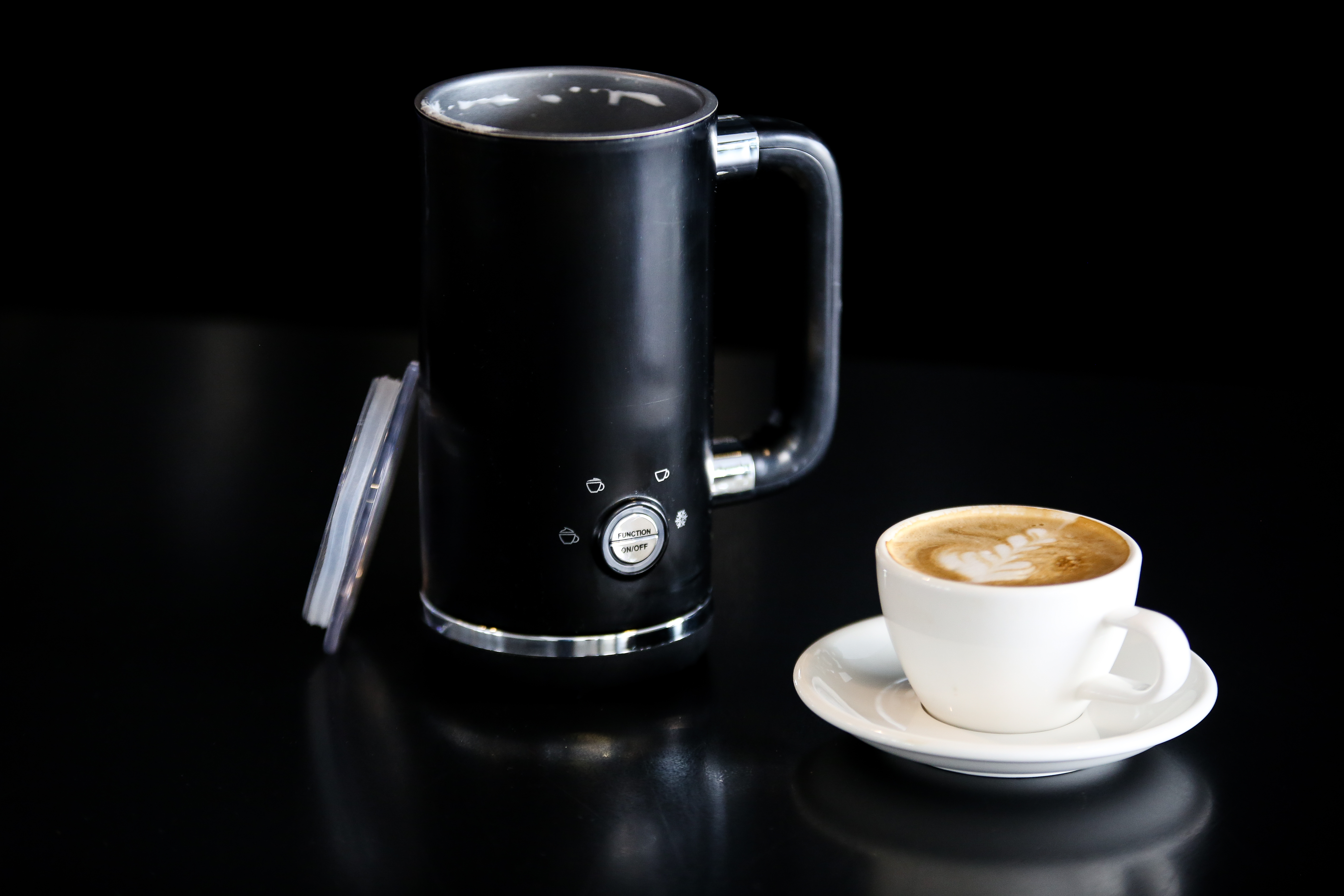 Schwarzer elektrischer Milchaufschäumer mit einer Tasse Latte und Untertasse auf schwarzer Oberfläche.