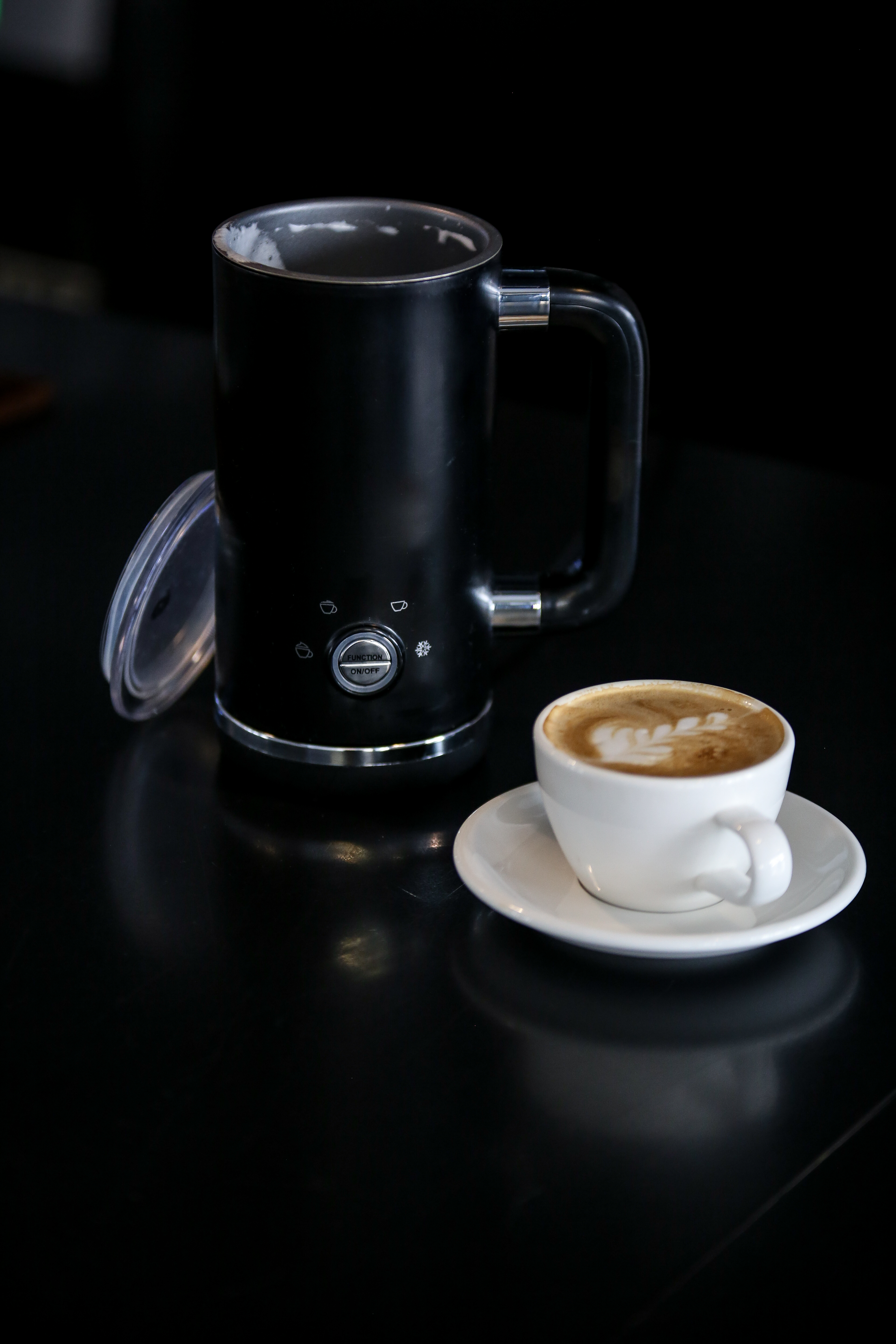 Schwarzer Milchaufschäumer mit einer Tasse Latte Art und Untertasse auf einem schwarzen Tisch.