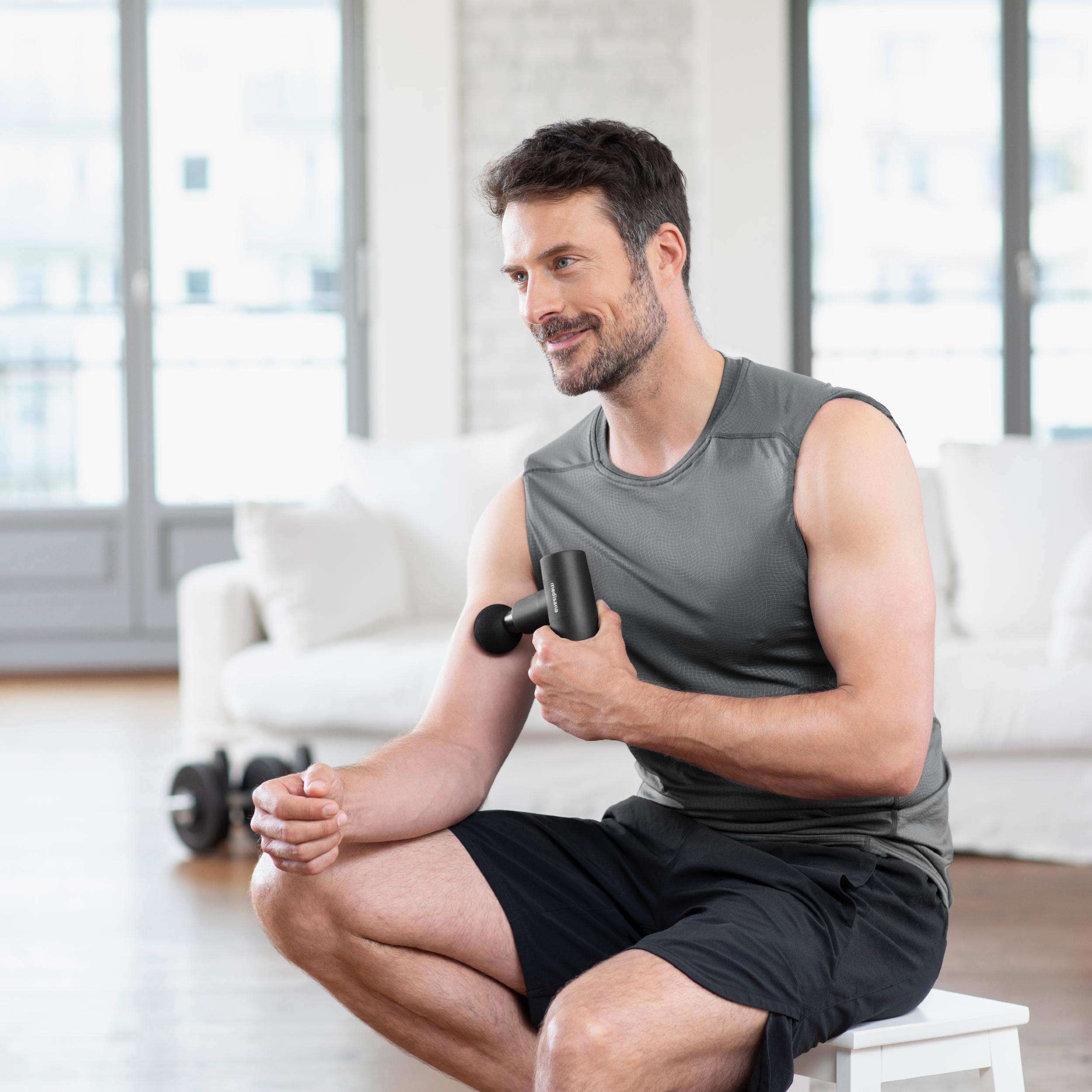 Ein Mann sitzt und benutzt eine Massagepistole an seinem Arm, lächelnd, mit Sofa im Hintergrund.