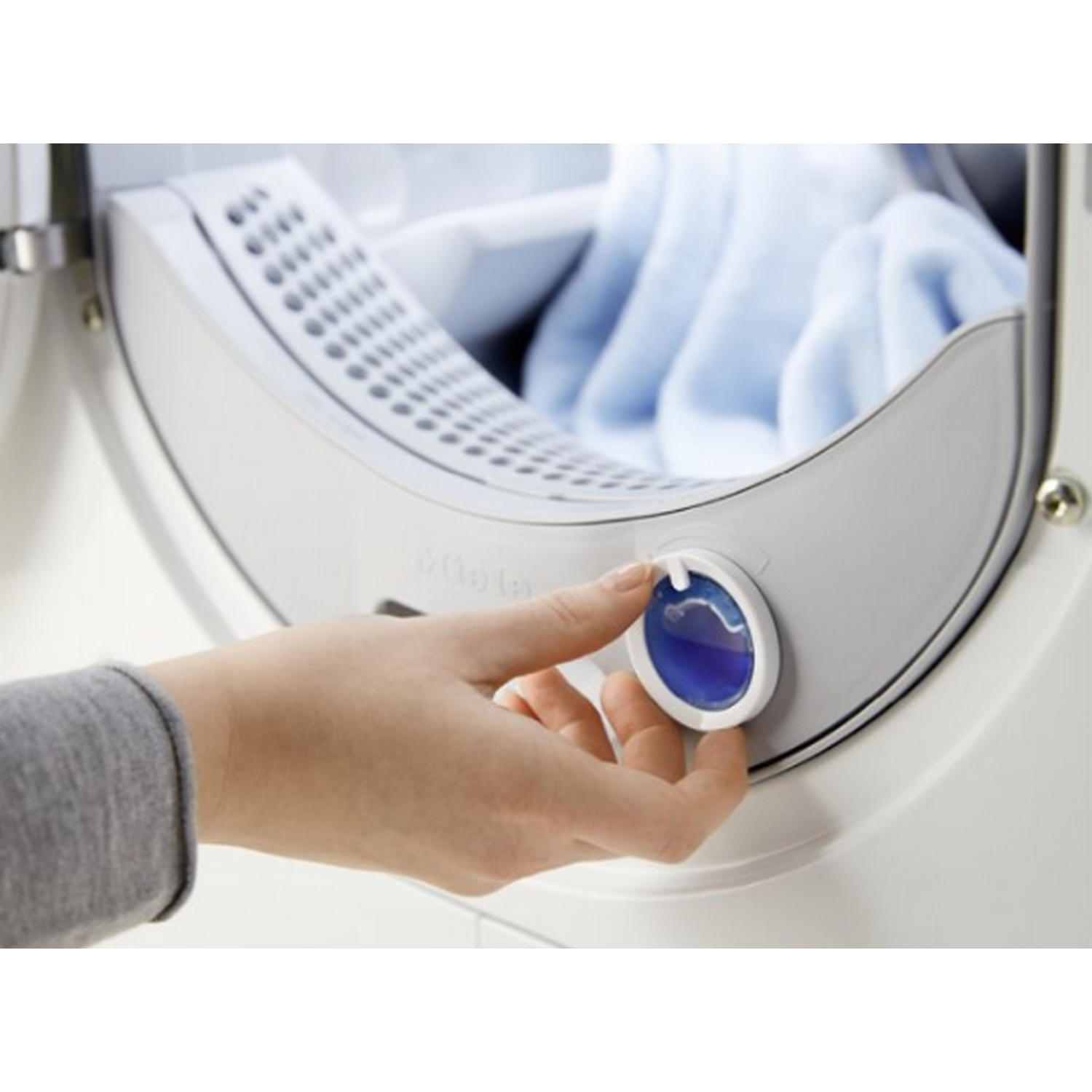 Una mano gira un dial azul en una lavadora blanca. Dentro, se ven prendas azules.
