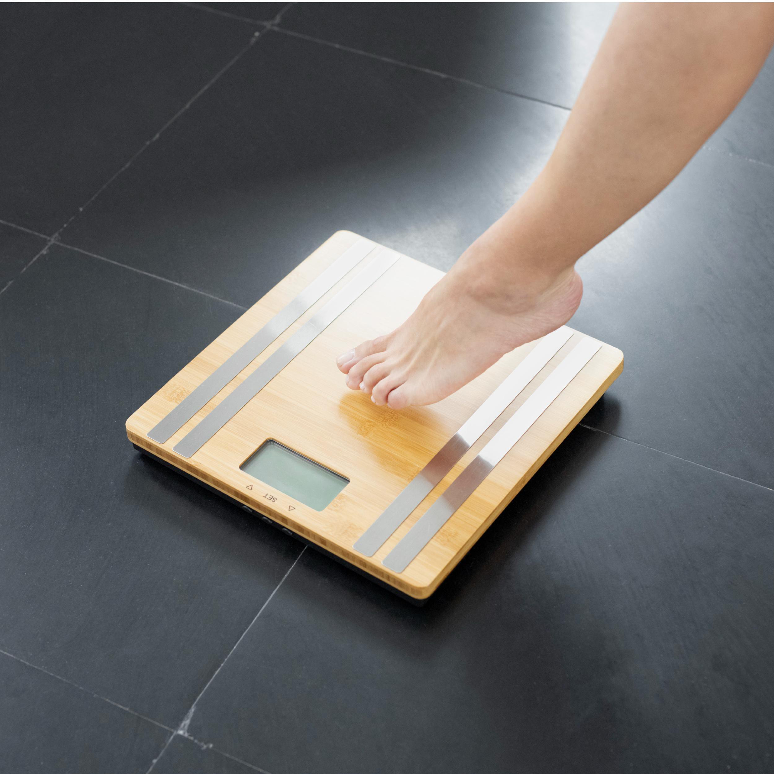 Der Fuß einer Person steht auf einer Holzwaage mit digitaler Anzeige. Die Waage steht auf einem schwarzen Fliesenboden.