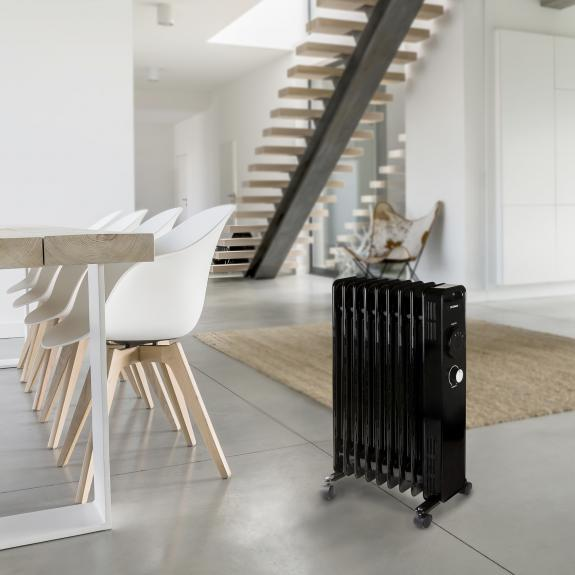 A black oil-filled radiator on wheels in a room. A white table and chairs are in the background.