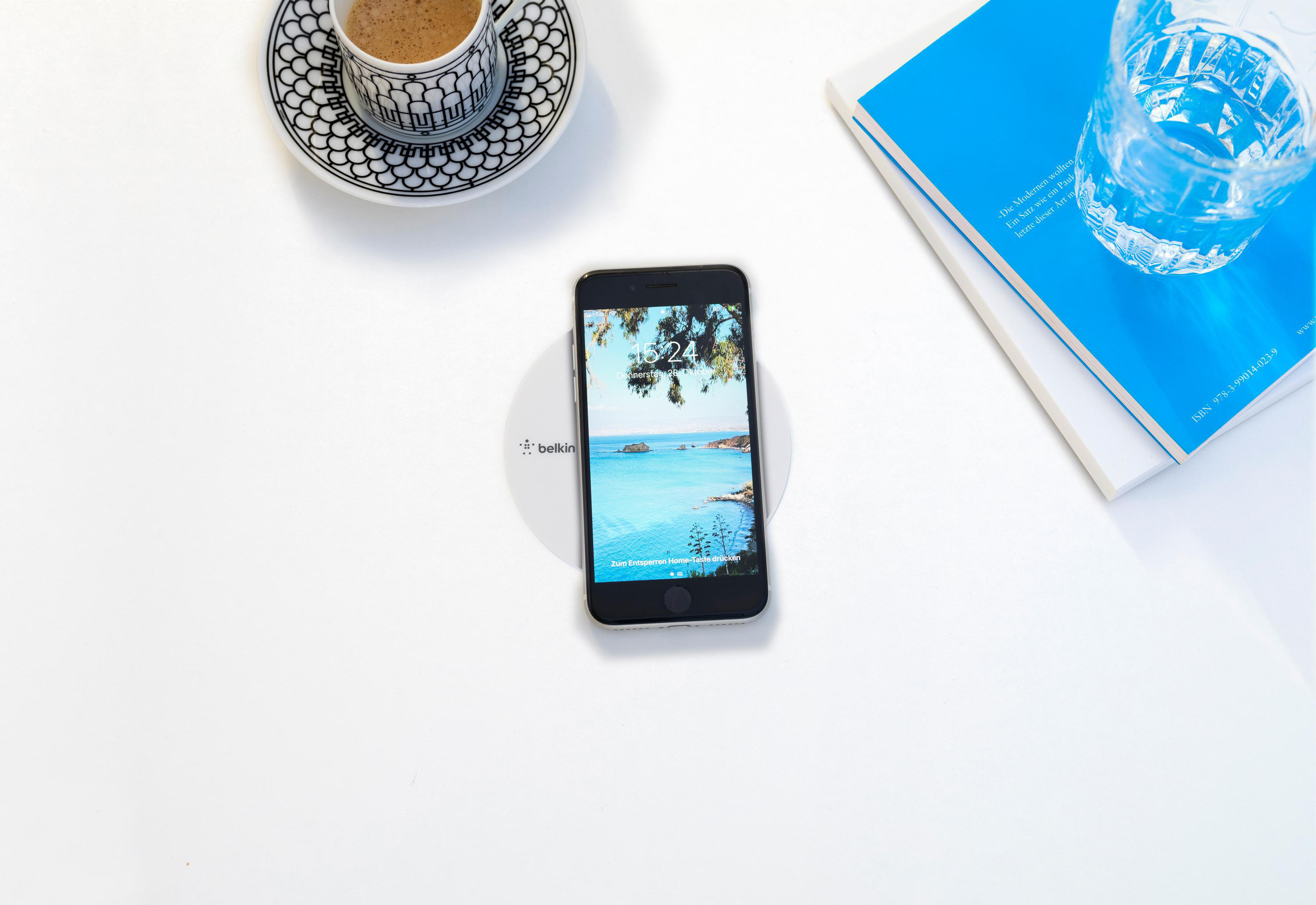 Telefon auf kabellosem Ladegerät, Kaffee, Glas Wasser und ein blaues Buch auf weißem Tisch.