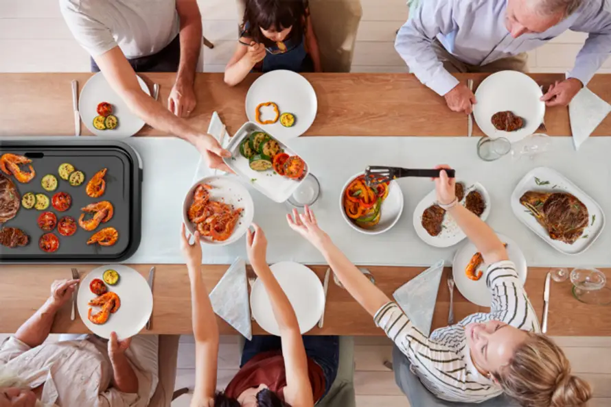 Une famille partageant de la nourriture autour d'une table, avec un grill à gauche et des assiettes de nourriture.