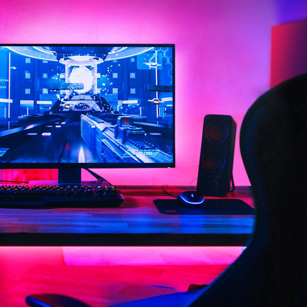 Gaming setup: monitor játékot jelenít meg, billentyűzet, egér, hangszórók és szék. Rózsaszín és kék fény.