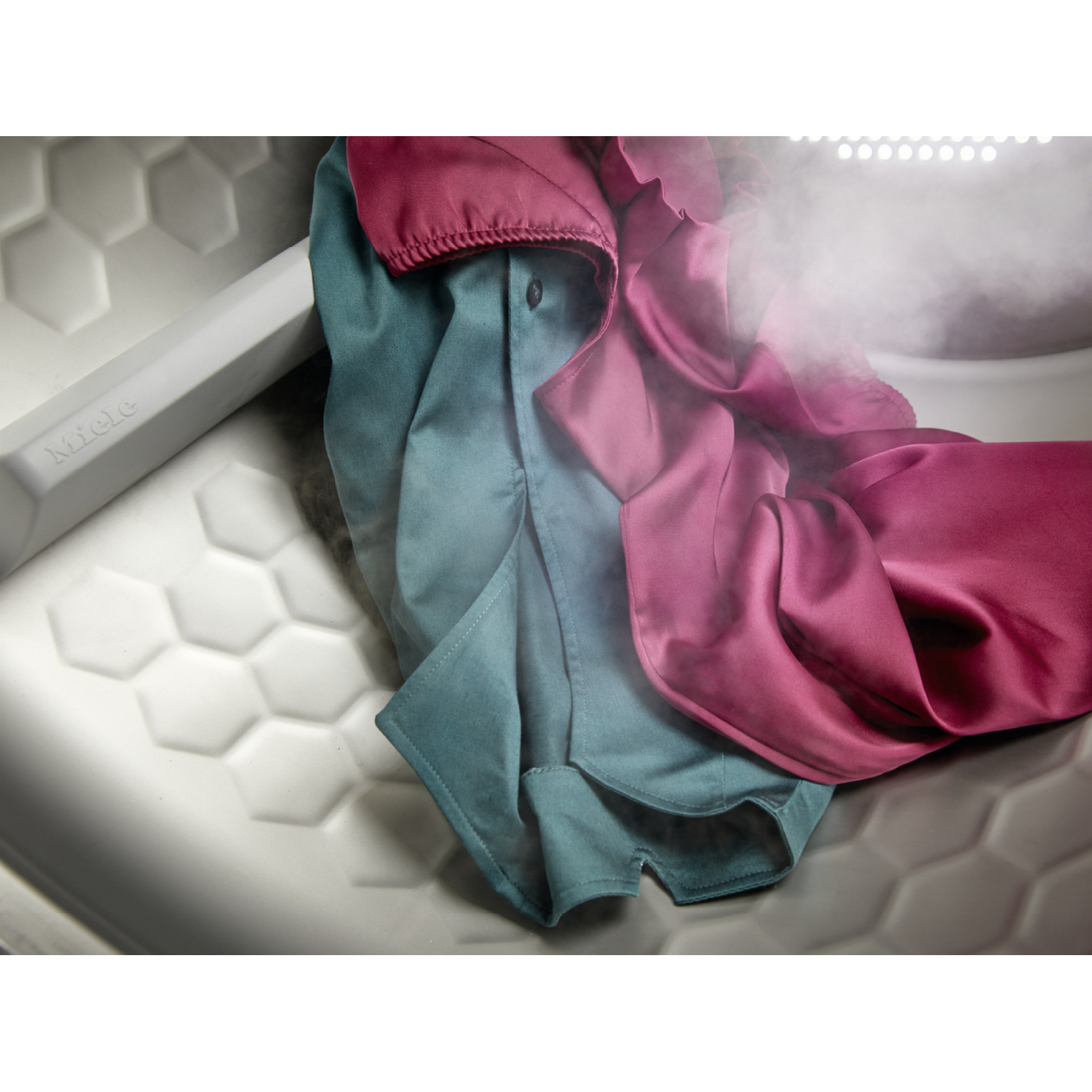 Dos camisas, una verde azulado, una rosa, en una lavadora. Vapor subiendo. Fondo blanco.