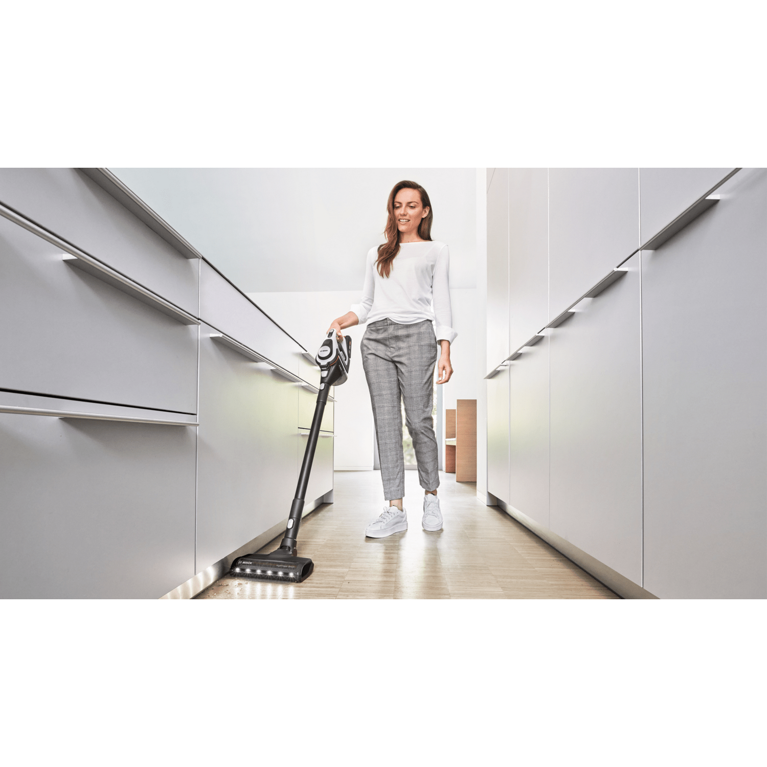 Mujer aspirando una cocina. Armarios blancos y suelo de madera al fondo.