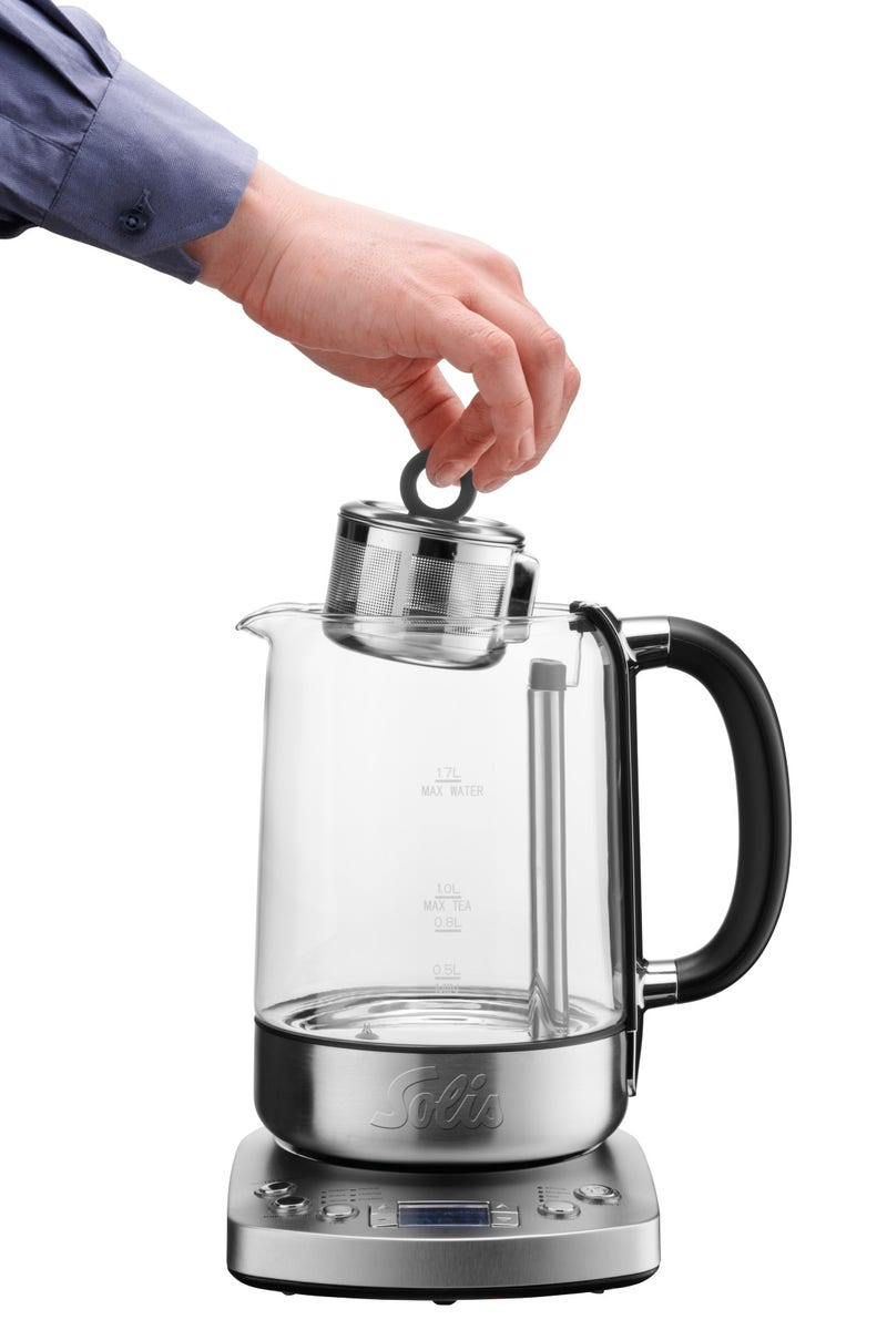 Main plaçant un infuseur à thé dans une bouilloire électrique en verre, sur fond blanc.