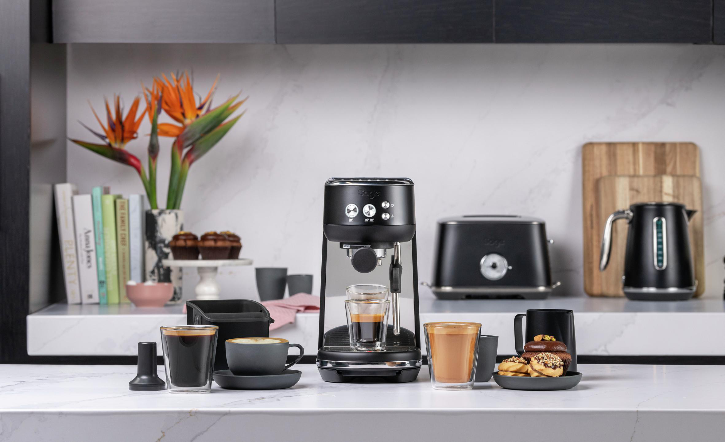 Küchentheke mit Espressomaschine, Kaffeezubehör und Gebäck, mit orangefarbenen Blumen und Büchern im Hintergrund.