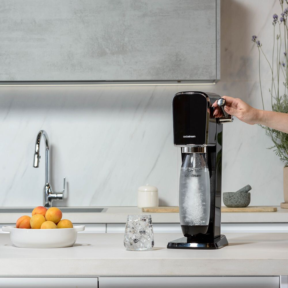 Machine SodaStream noire, verre avec glace et bol d'abricots sur comptoir blanc.