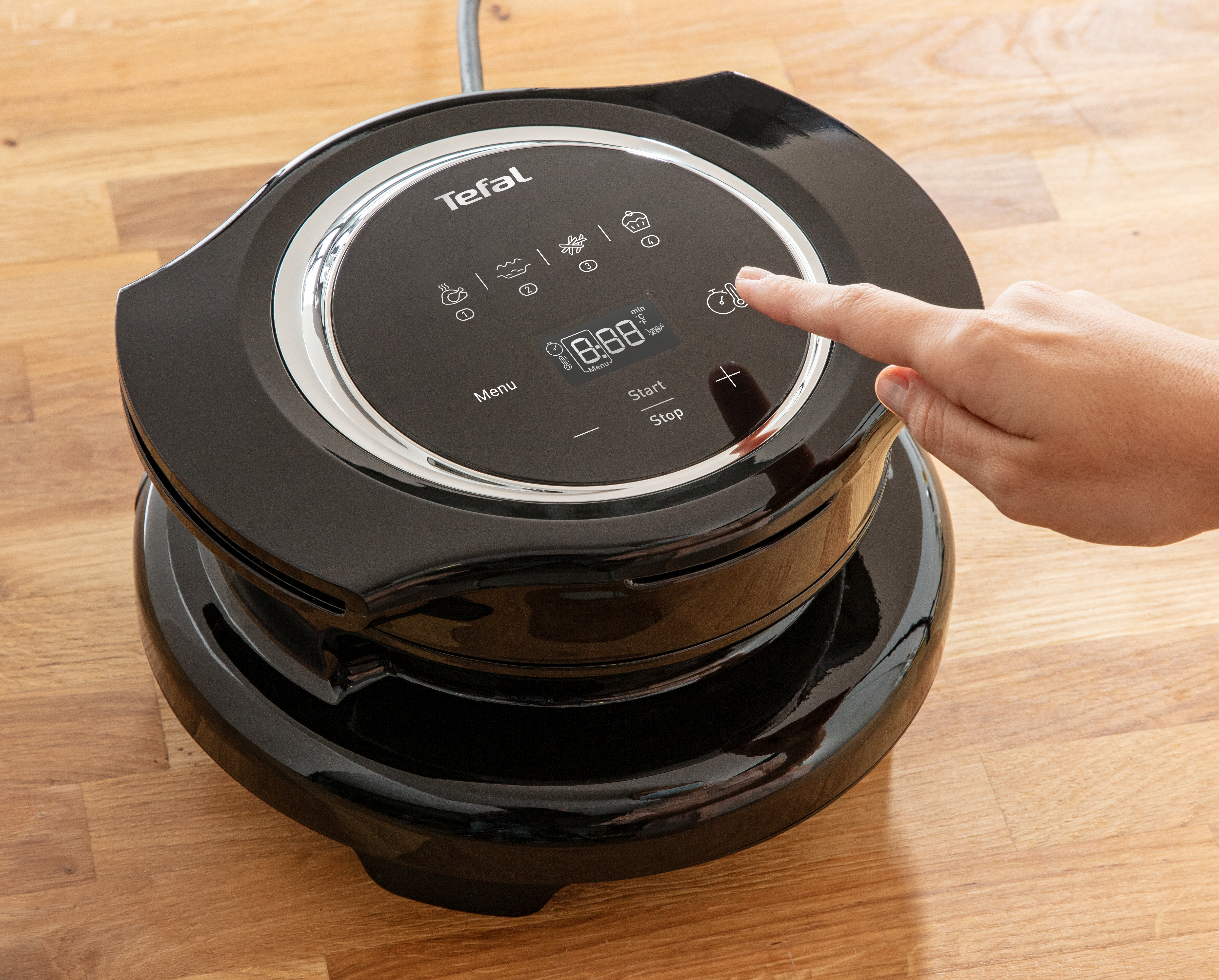 Fekete Tefal készülék érintőképernyővel, egy kéz érinti a képernyőt, fa felületen, közelkép.