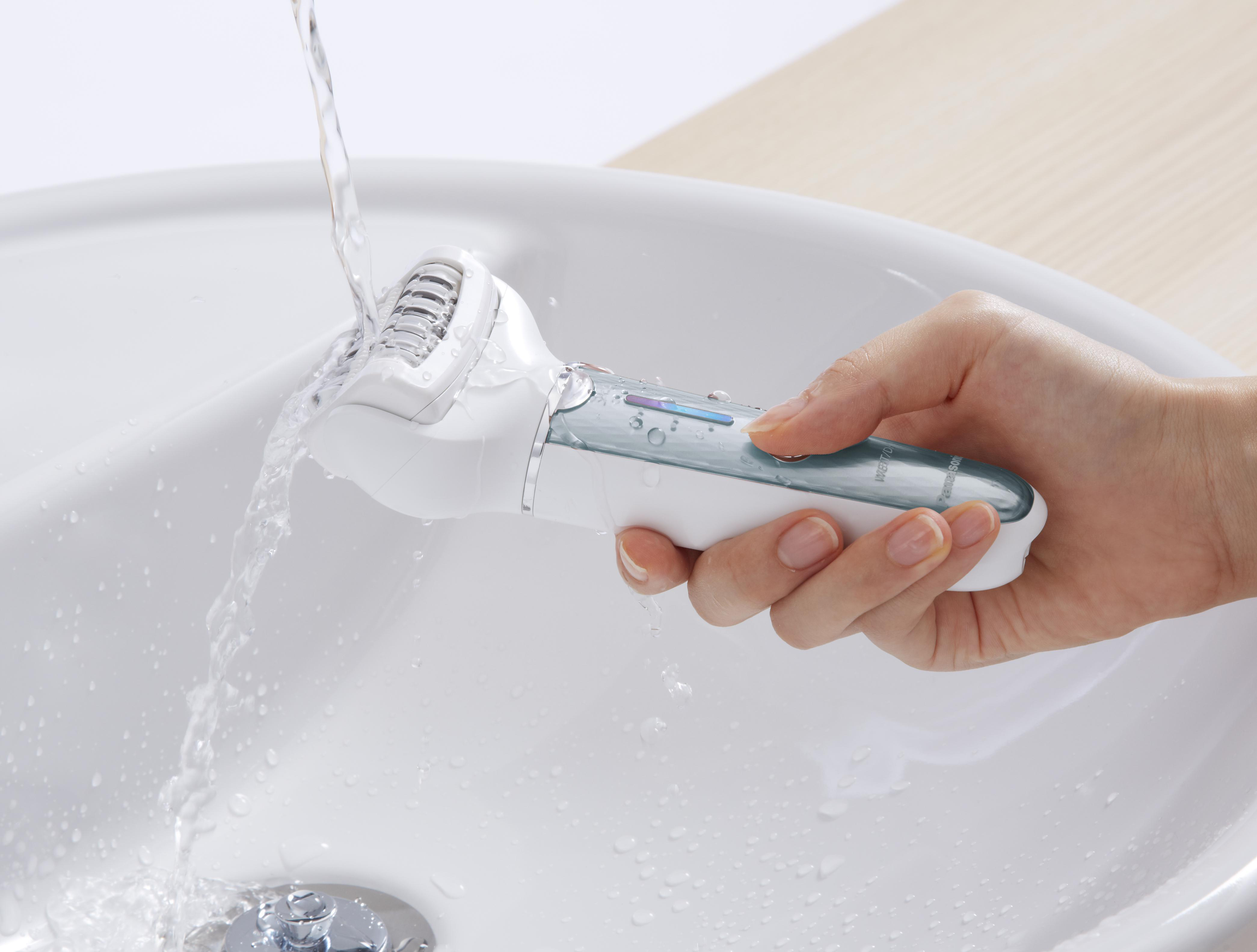Die Hand einer Frau hält einen weißen Elektrorasierer unter fließendem Wasser in einem weißen Waschbecken.