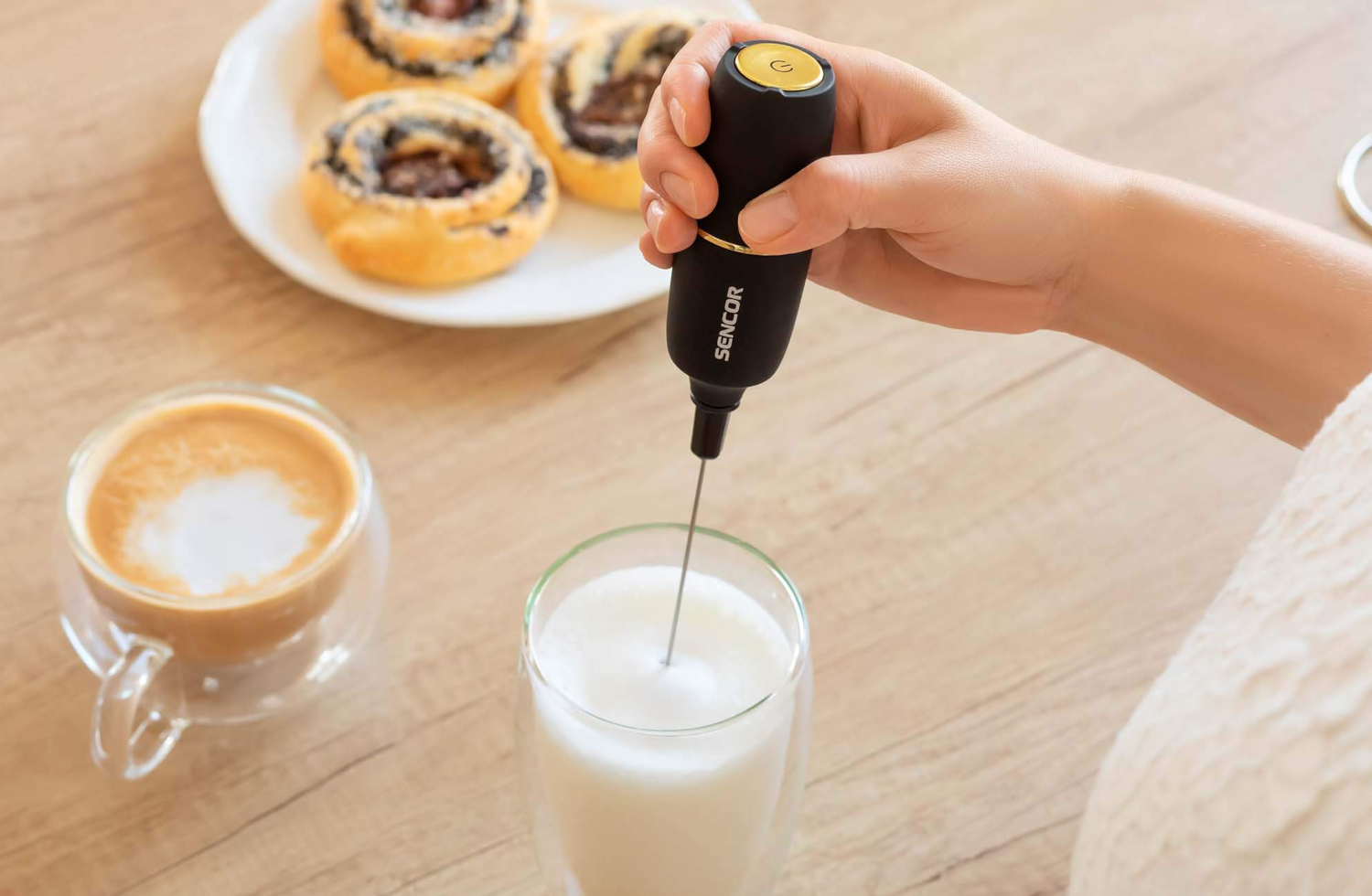 Egy kéz egy fekete tejhabosítót tart, tejet habosítva egy pohárban. Kávé és sütemények a háttérben.
