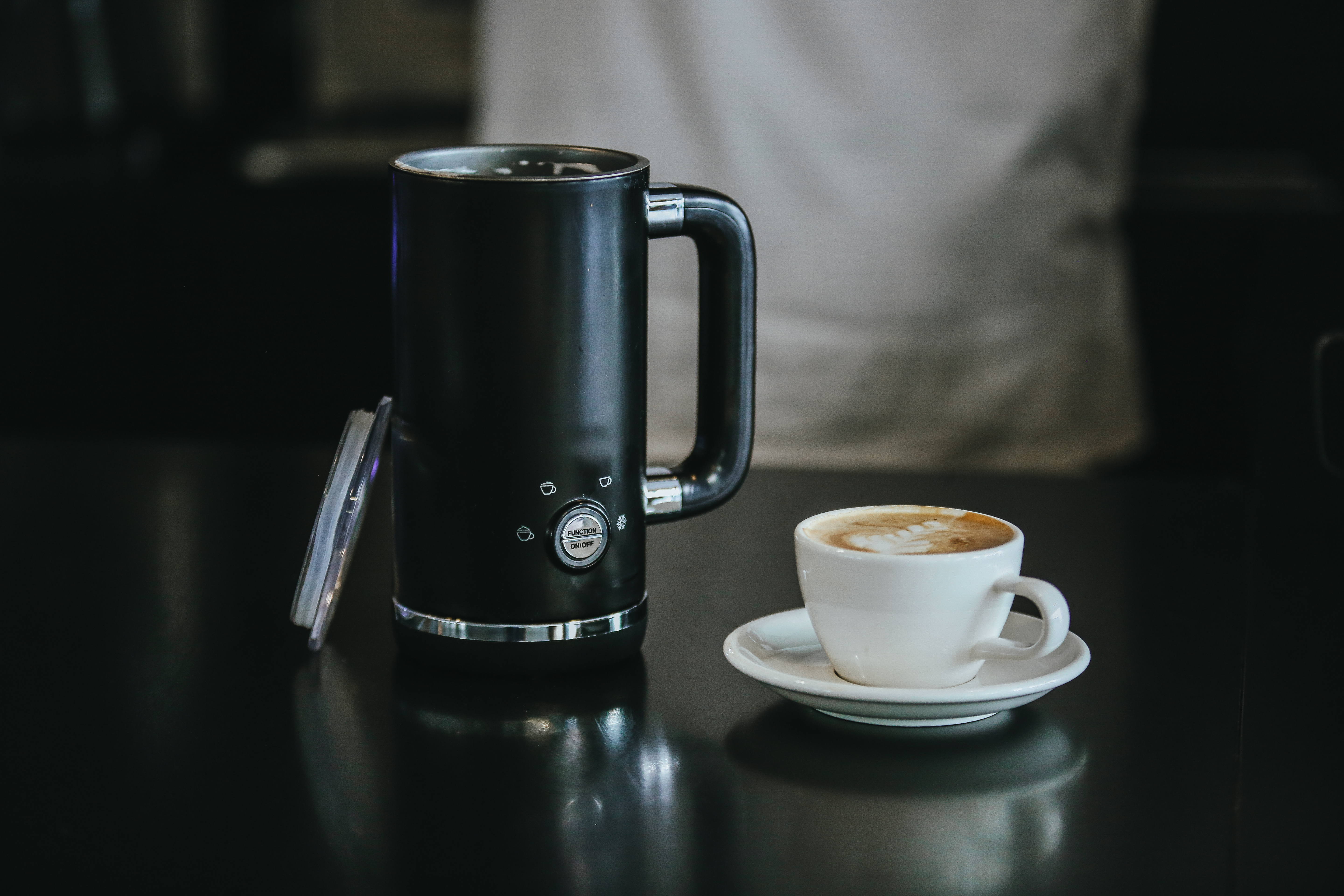 Schwarzer Milchaufschäumer und eine Tasse Kaffee auf dunkler Oberfläche. Person im Hintergrund.