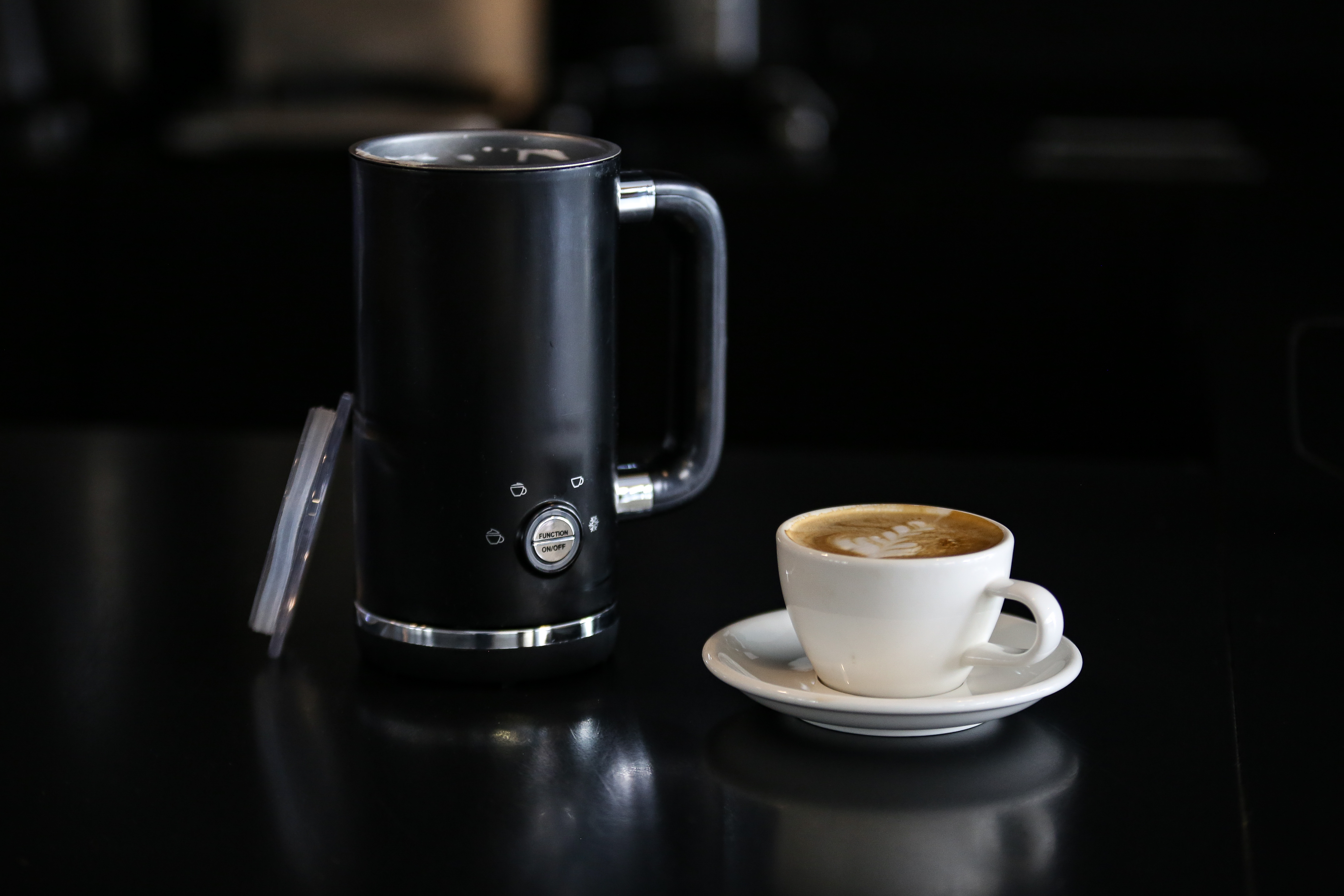 Schwarzer Milchaufschäumer mit einer Tasse Kaffee auf reflektierender schwarzer Oberfläche.