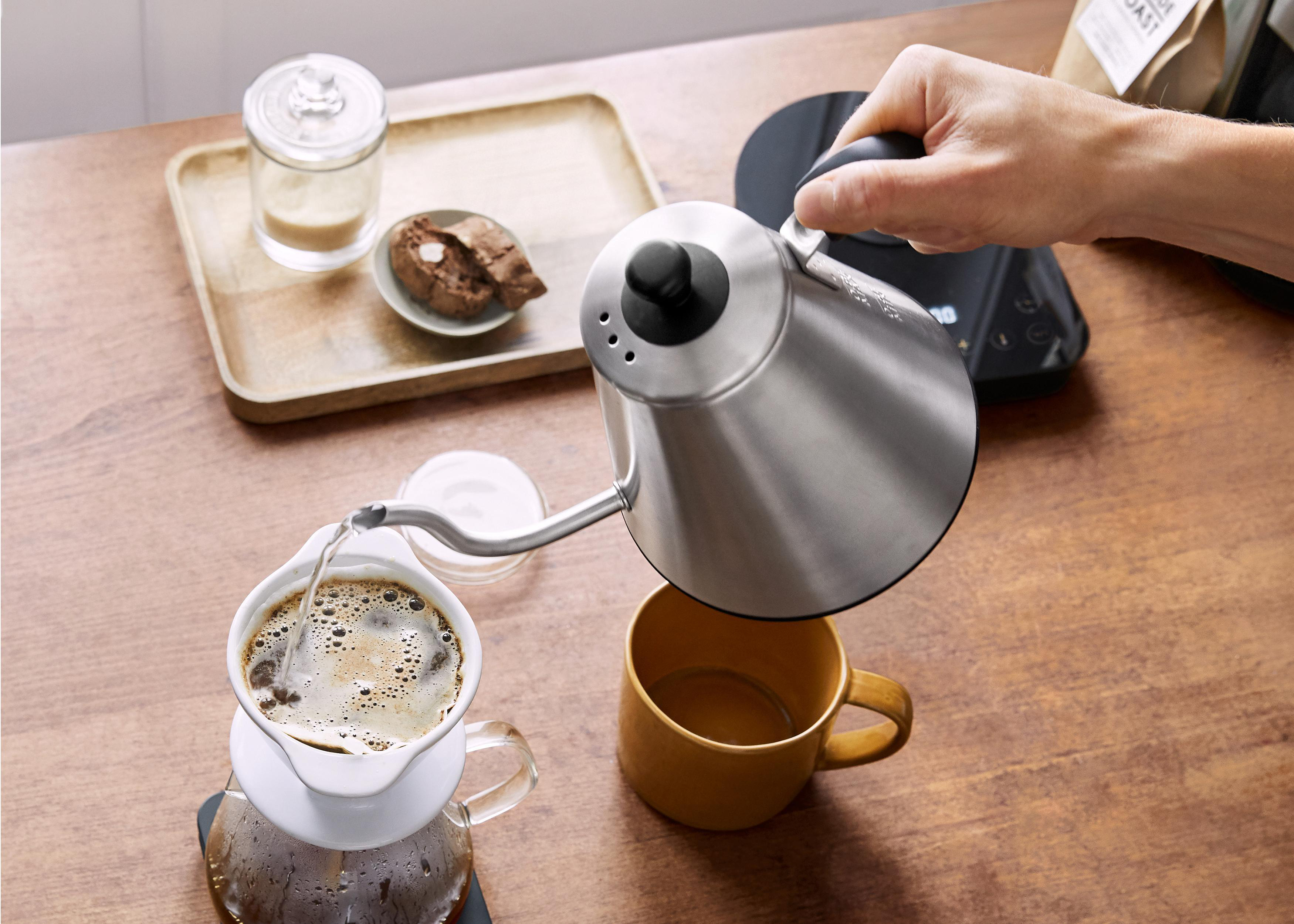 Eine Person gießt Wasser aus einem silbernen Wasserkocher in einen weißen Kaffeefilter auf einer Tasse. Holztisch.