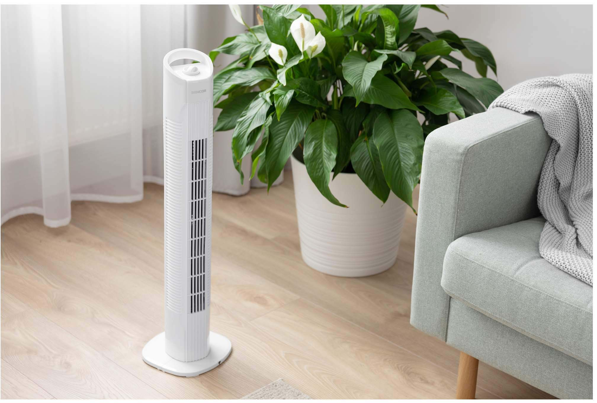 Fehér toronyventilátor egy szobában növényekkel és szürke kanapéval.
