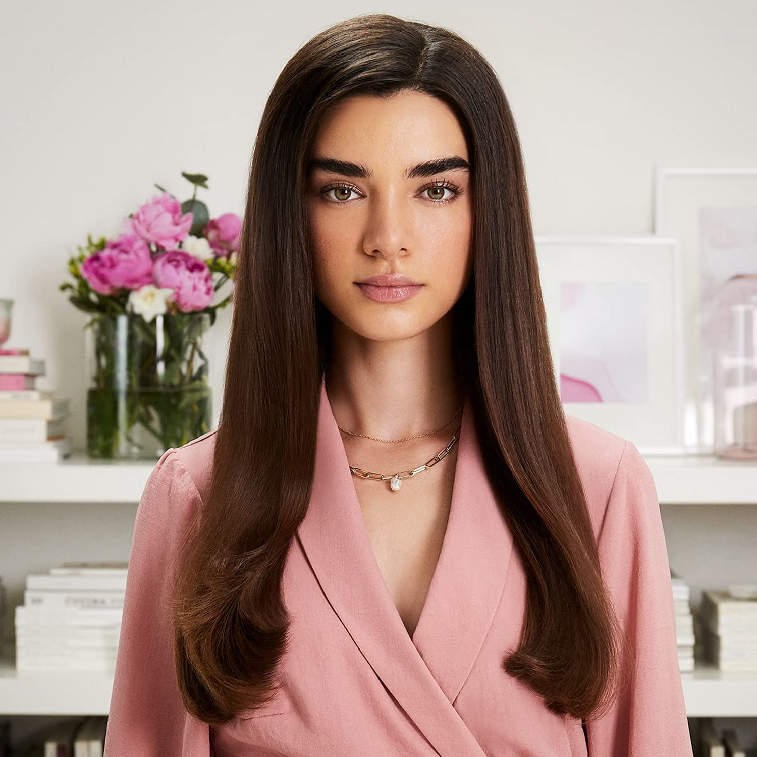 Una mujer con cabello largo y liso castaño que lleva una blazer rosa. Hay flores en el fondo.