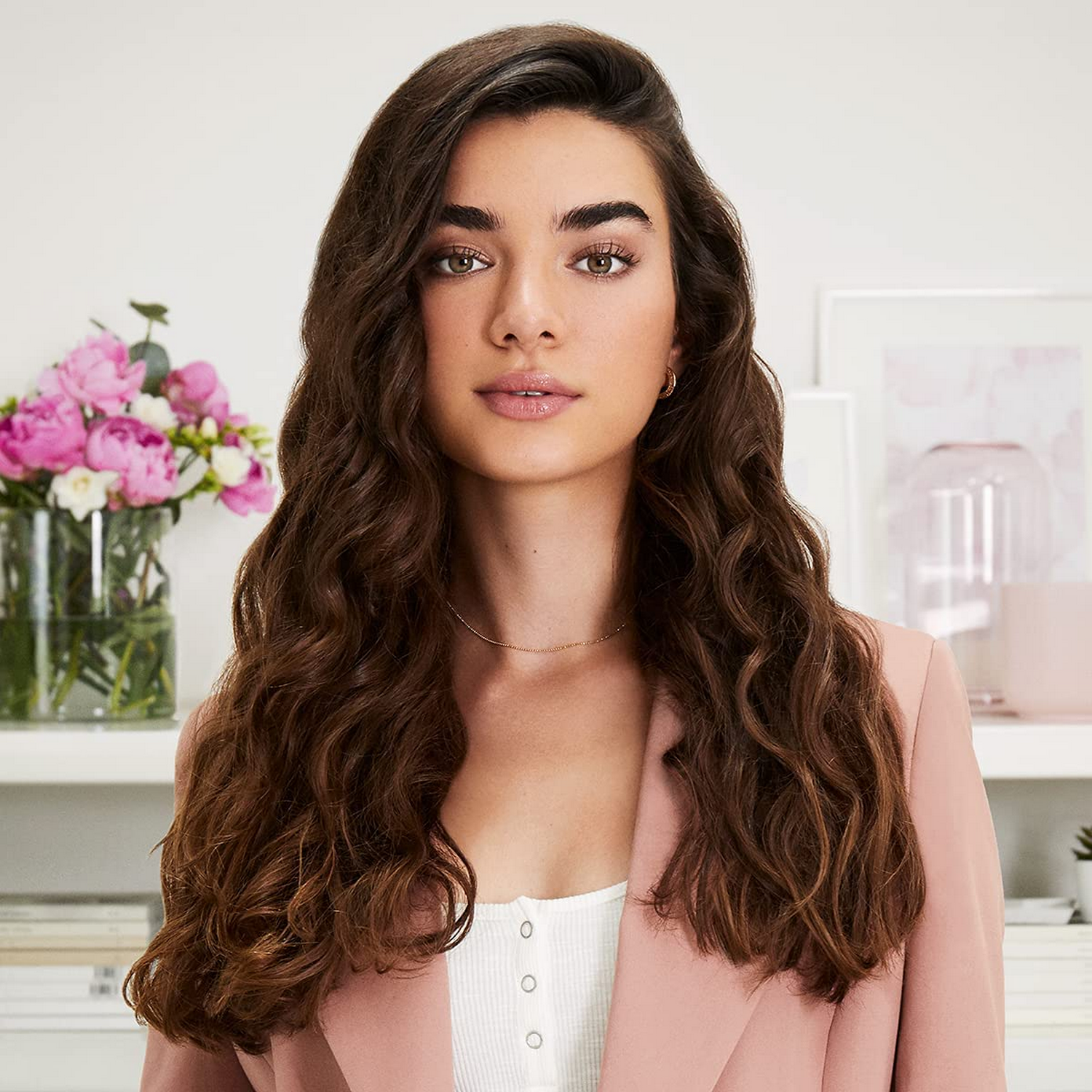 Mujer con largo cabello castaño ondulado, blazer rosa, top blanco y flores al fondo.