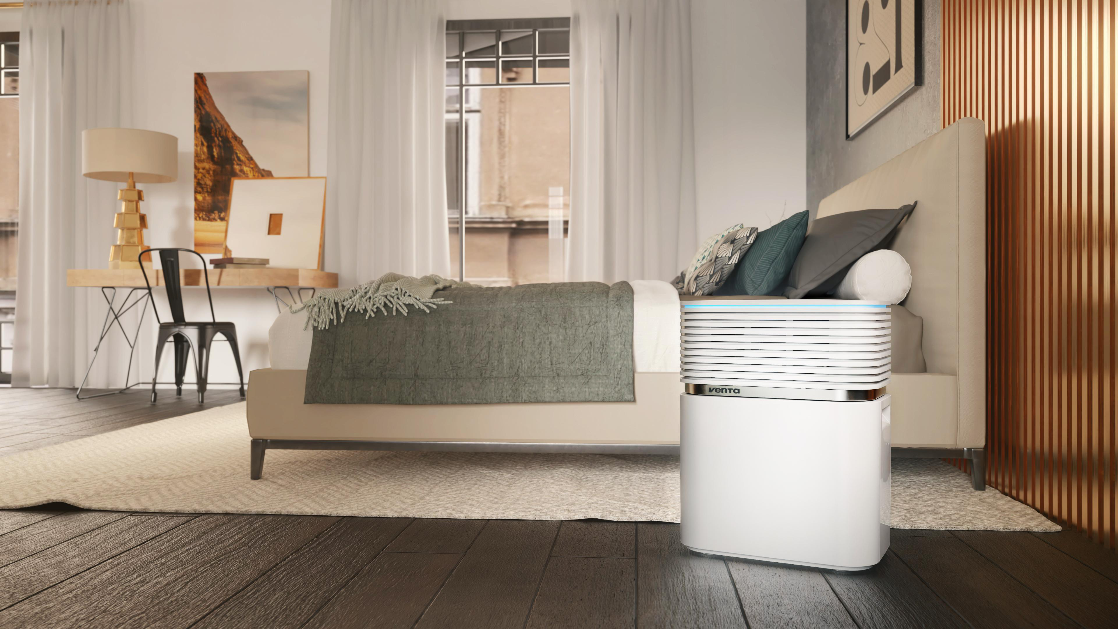 Schlafzimmer mit Bett, Luftreiniger und Kunstwerk. Das Bett hat eine graue Decke. Der Luftreiniger ist weiß.
