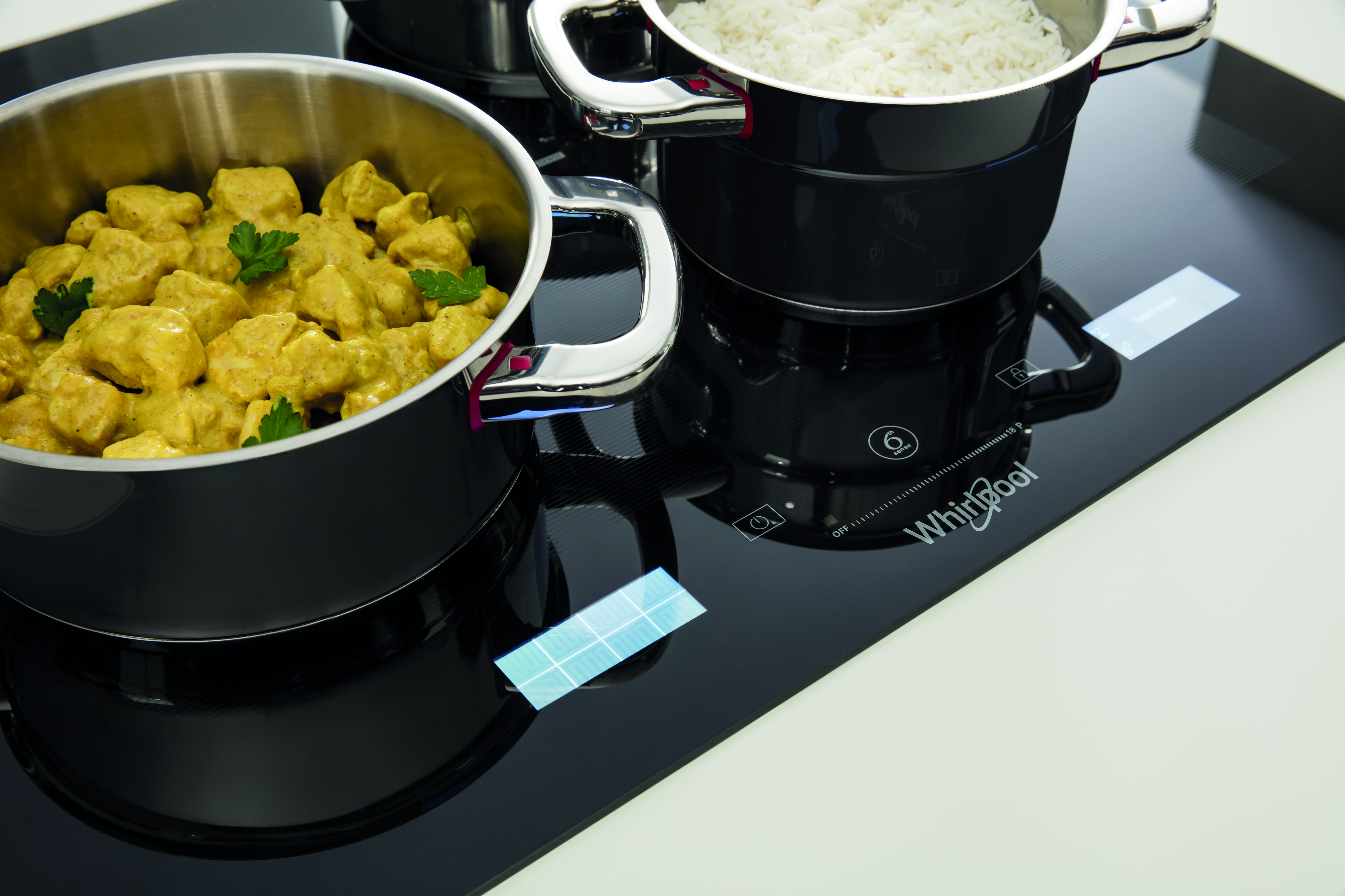 Főzőedények egy fekete főzőlapon. Az egyik edényben curry, a másikban fehér rizs van. Fehér felület.