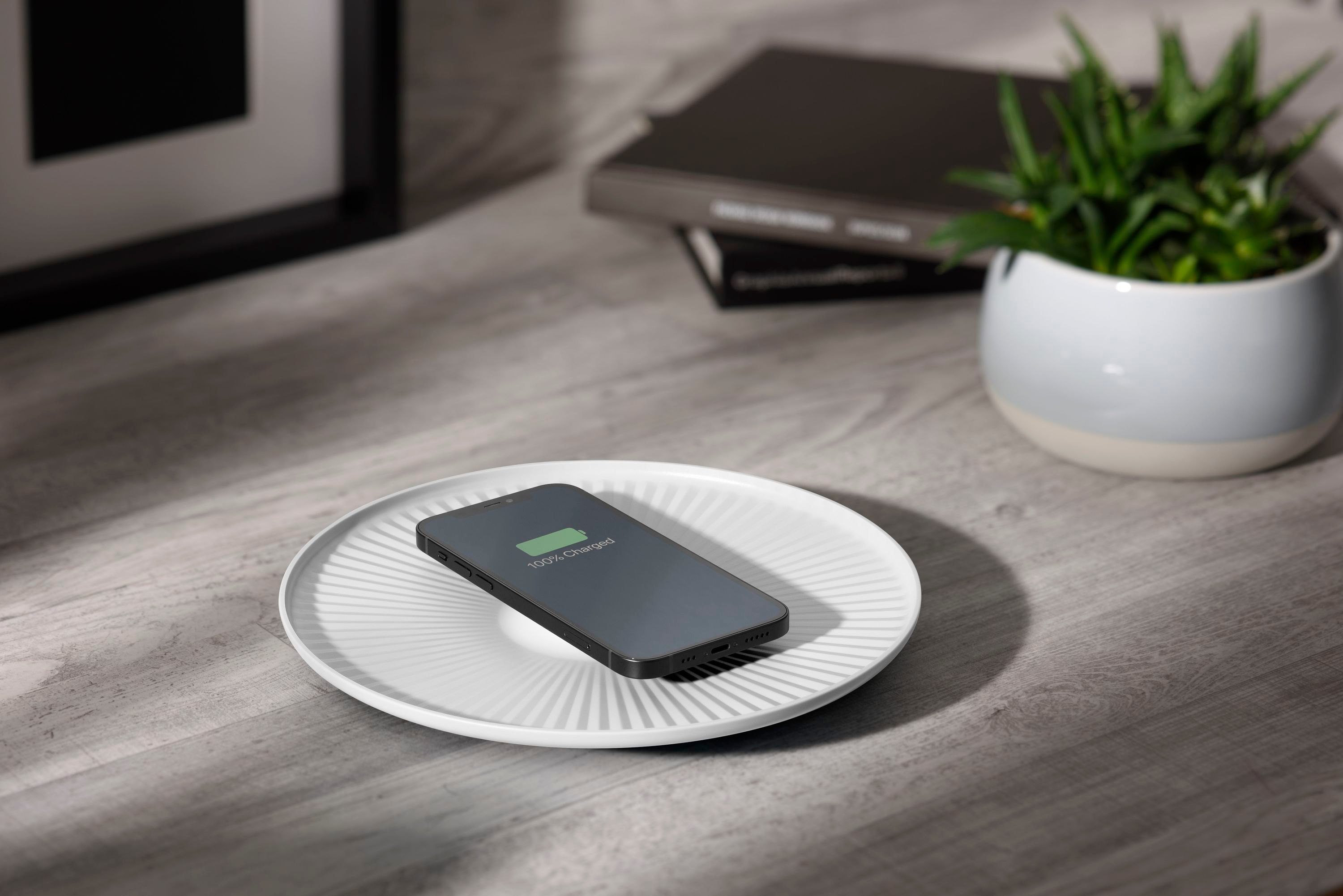 Un téléphone se chargeant sur une assiette blanche, sur un bureau gris, avec des plantes et des livres.