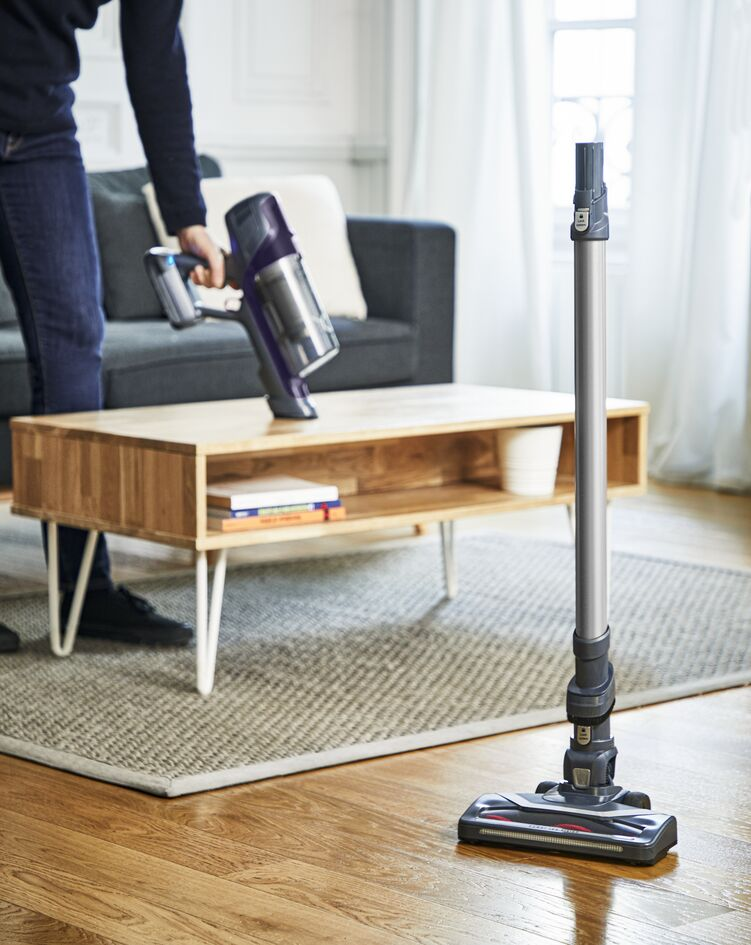 Persoon stofzuigt een houten vloer met een steelstofzuiger. Tafel en bank op de achtergrond.