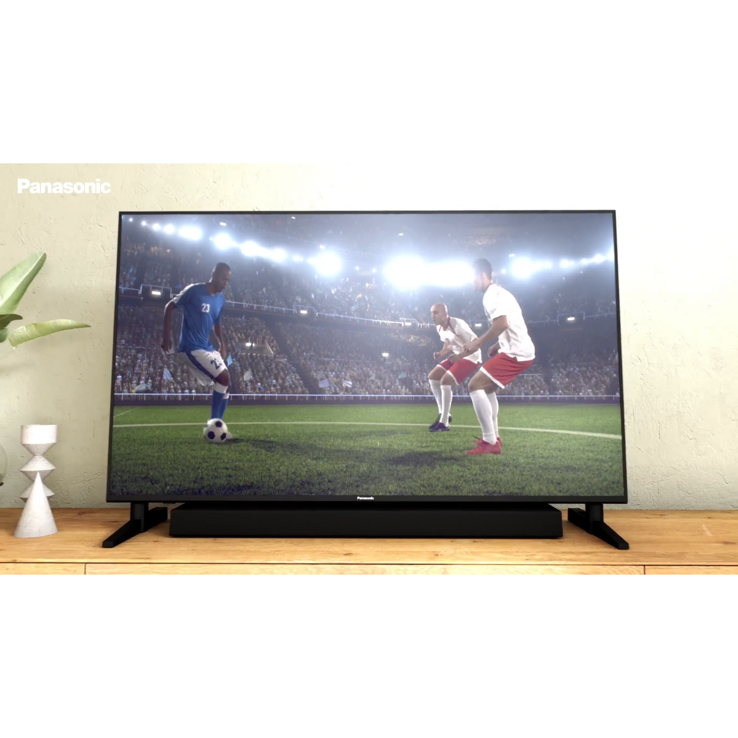 Un televisor muestra un partido de fútbol, jugadores en campo verde, barra de sonido abajo.