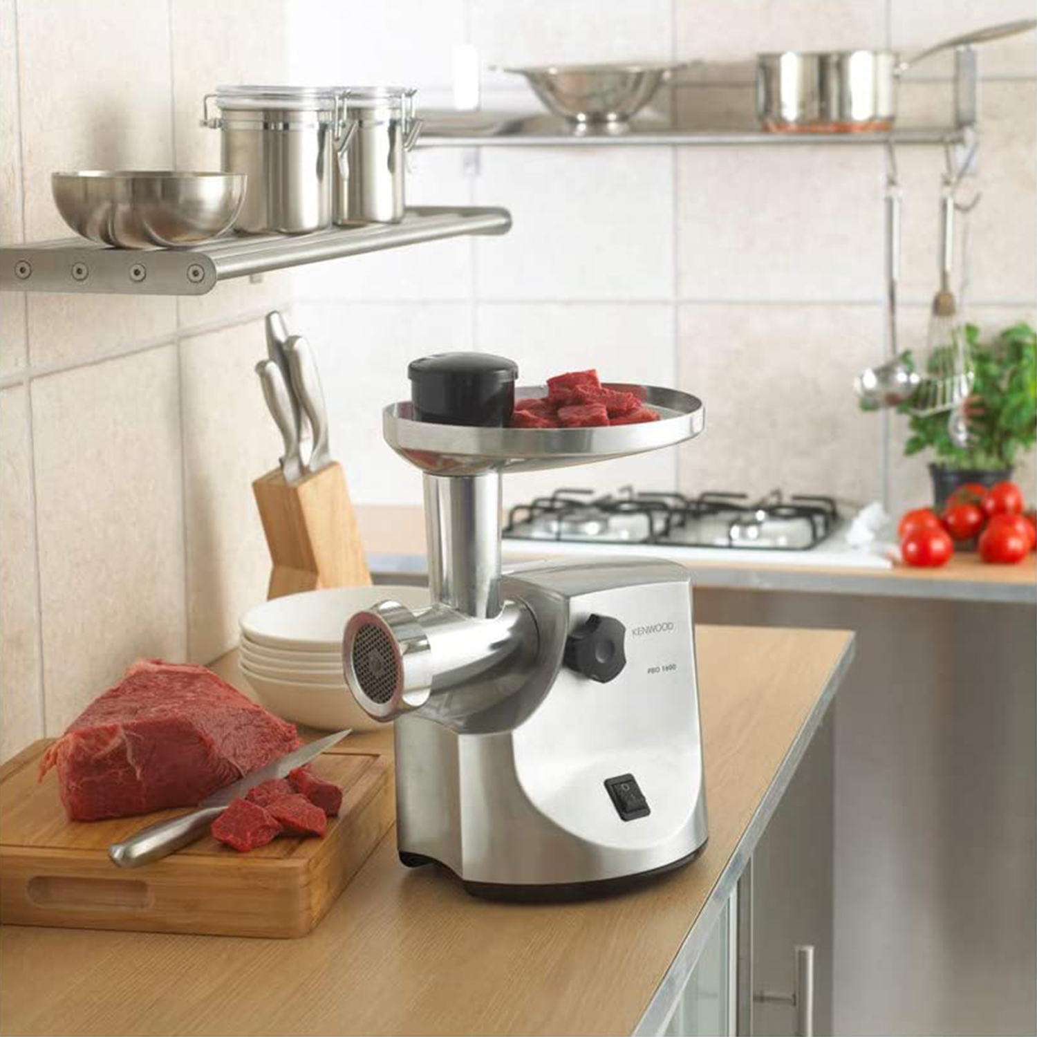 Tritacarne in acciaio inox con carne su tagliere e cucina sullo sfondo.