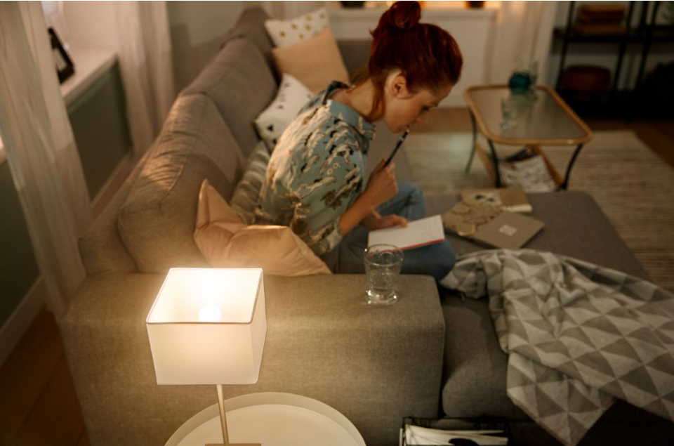 Femme sur canapé gris écrivant dans un cahier, lampe et verre d'eau.