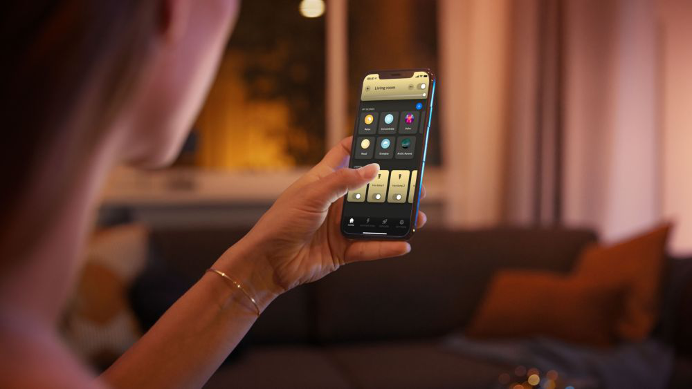 Femme utilisant un téléphone pour contrôler les lumières d'une pièce. Le téléphone affiche une interface d'application domotique.