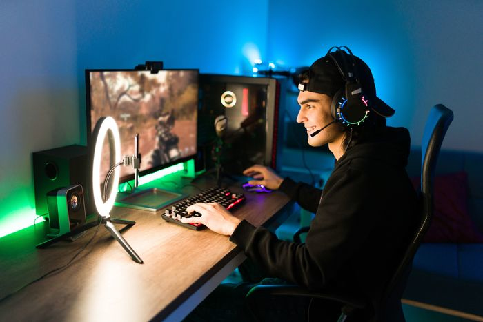 Gamer wearing headphones, playing at a desk with a monitor and ring light. Blue and purple lights.