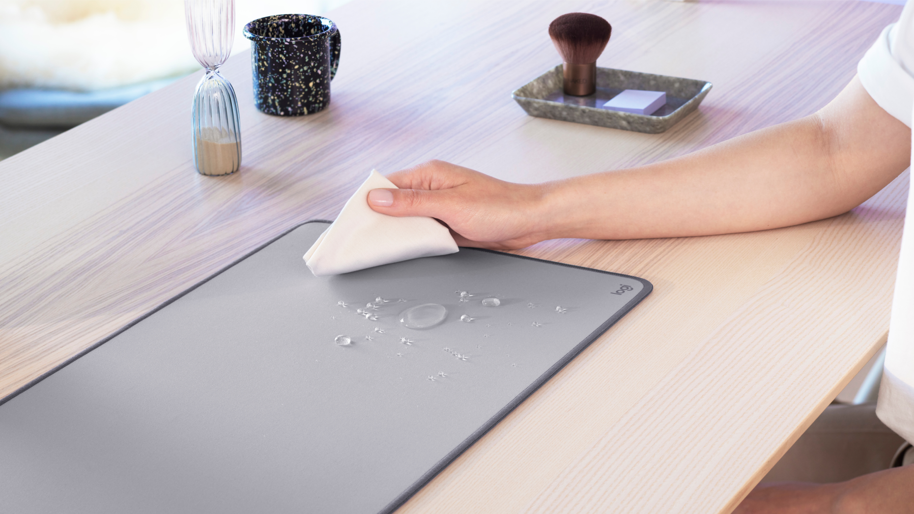 Une main essuyant un tapis de souris gris clair sur une table en bois, avec des gouttes d'eau et des accessoires en arrière-plan.