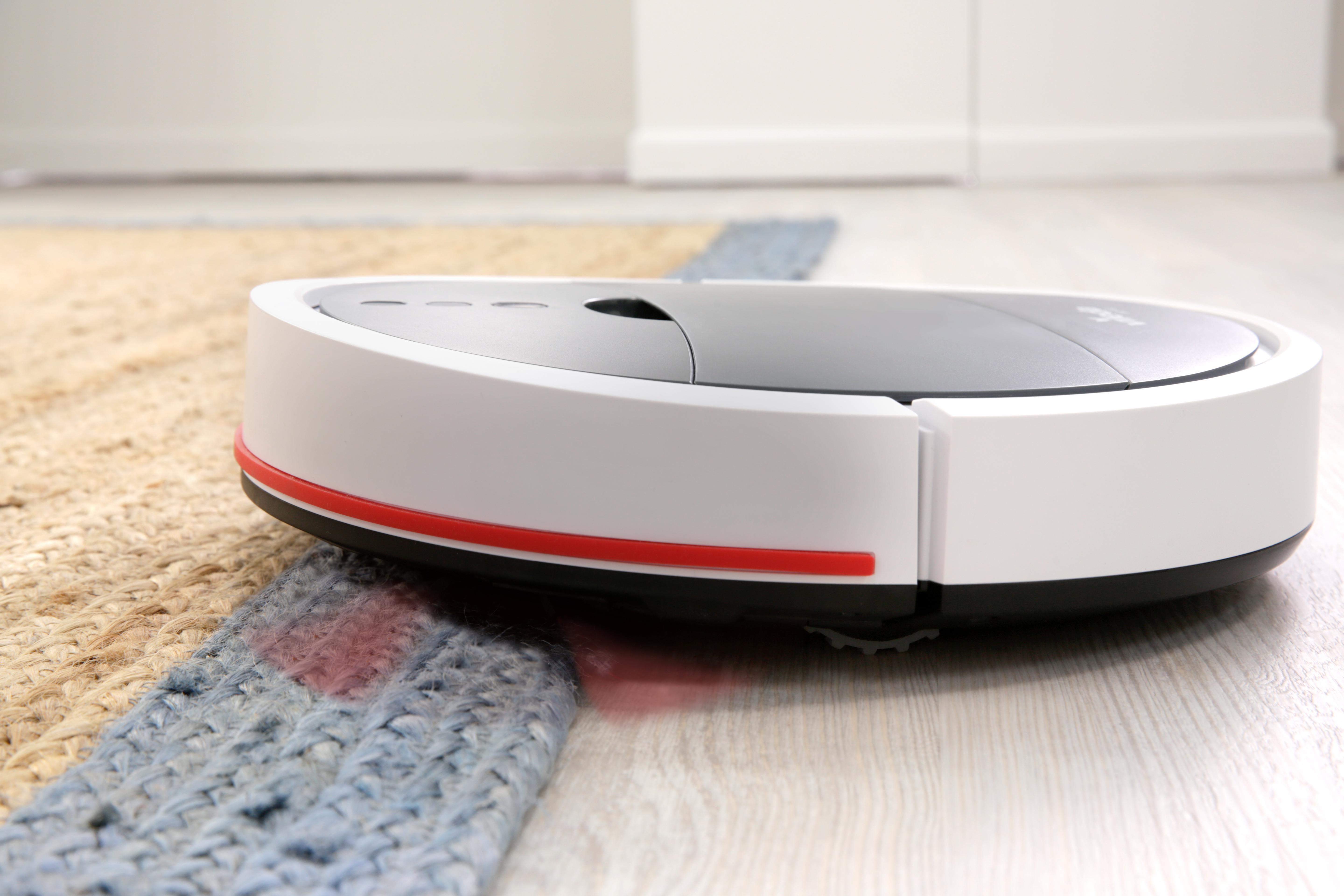 Roboter-Staubsauger auf einem Teppich, neben Holzboden, mit rotem Streifen und schwarzem Deckel.