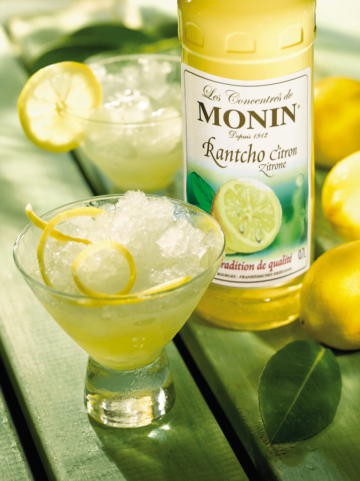 Cocktail mit Zitrone und Eis. Monin-Sirup-Flasche im Hintergrund. Grüner Hintergrund.
