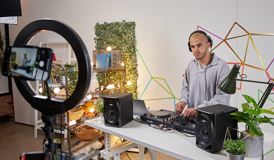 Uomo DJ con telefono che registra, circondato da attrezzature e vegetazione.
