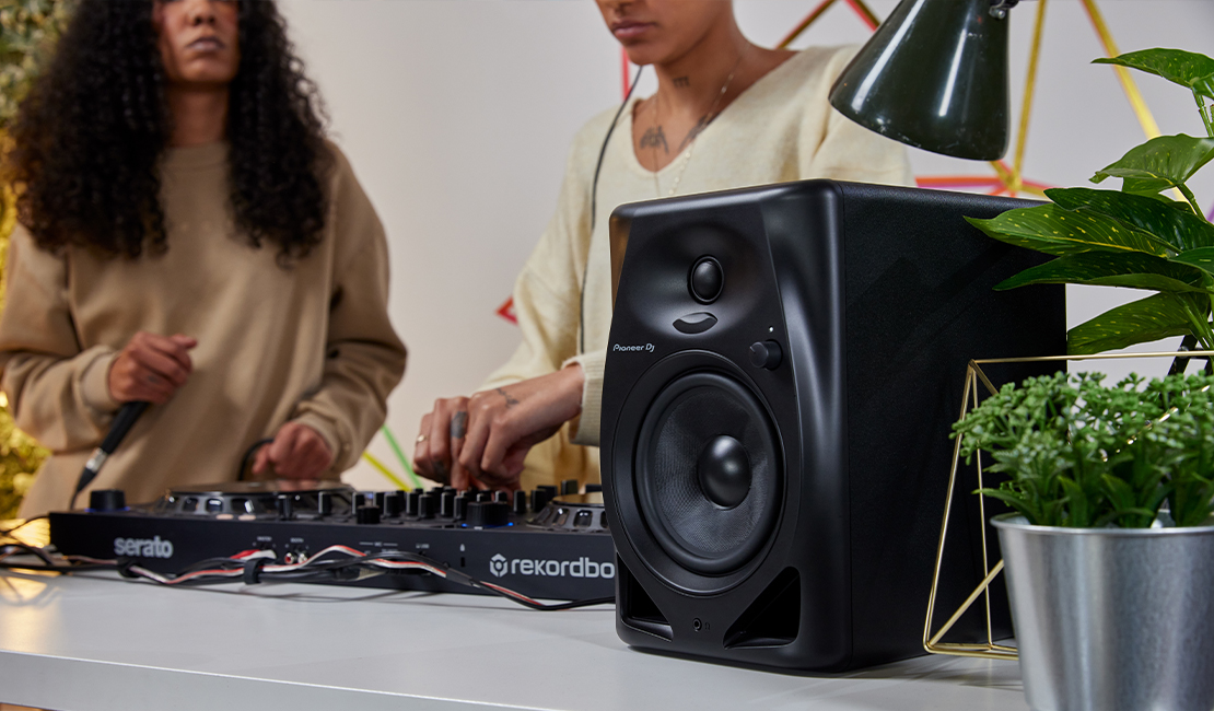 Due persone DJing con un mixer e altoparlanti su un tavolo bianco, una pianta sullo sfondo.