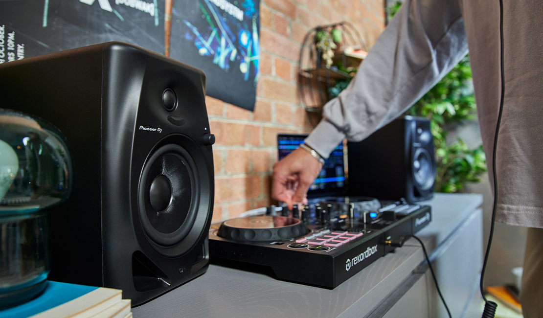 Una persona regola un mixer per DJ, con altoparlanti in primo piano. Si vede un muro di mattoni.