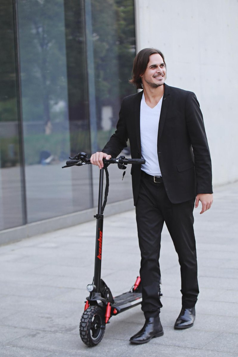 Homme en costume noir avec un scooter noir. Il sourit, debout sur un trottoir à côté d'un bâtiment.