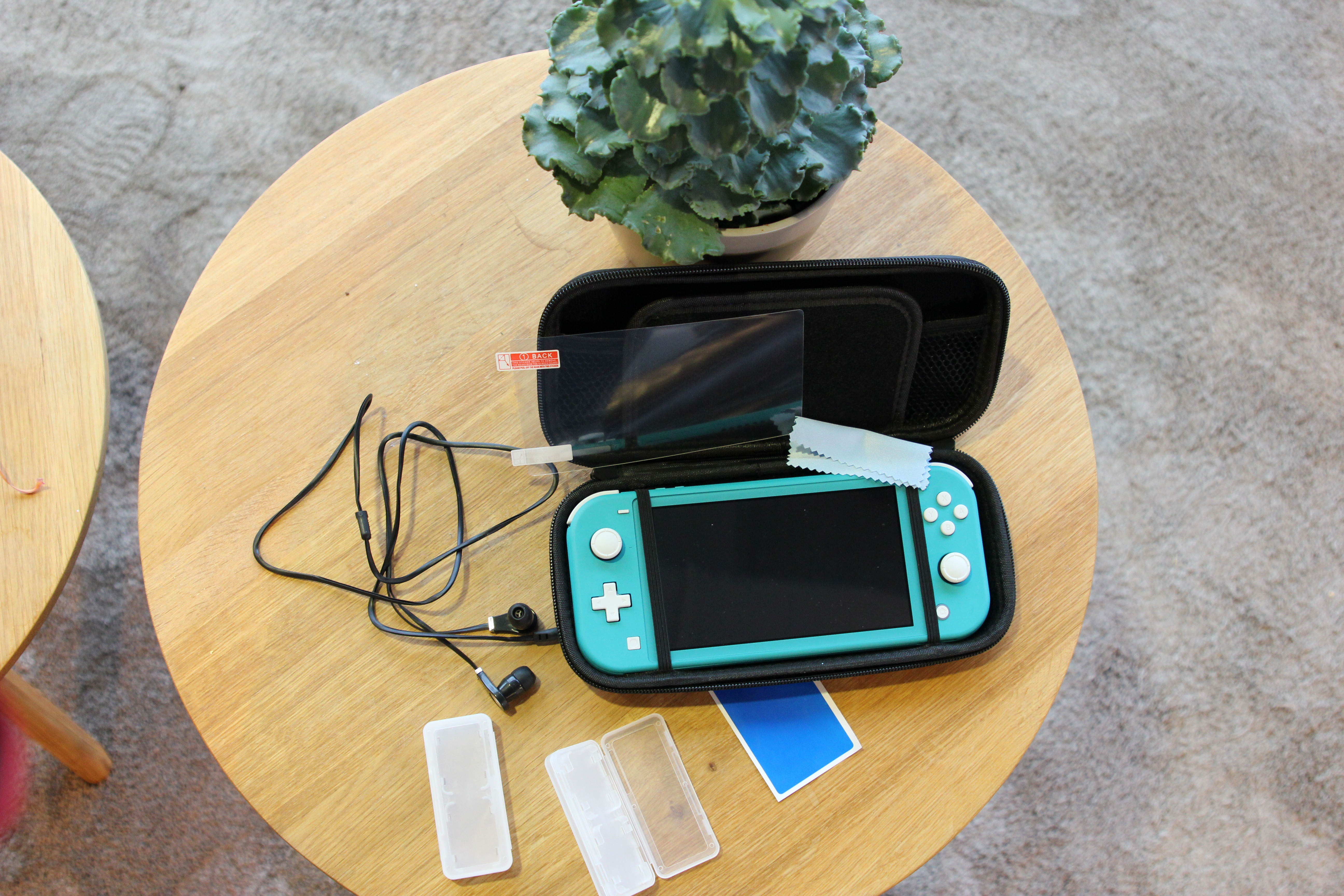 A teal Nintendo Switch Lite, case, screen protector, and accessories are on a round wooden table with a plant.
