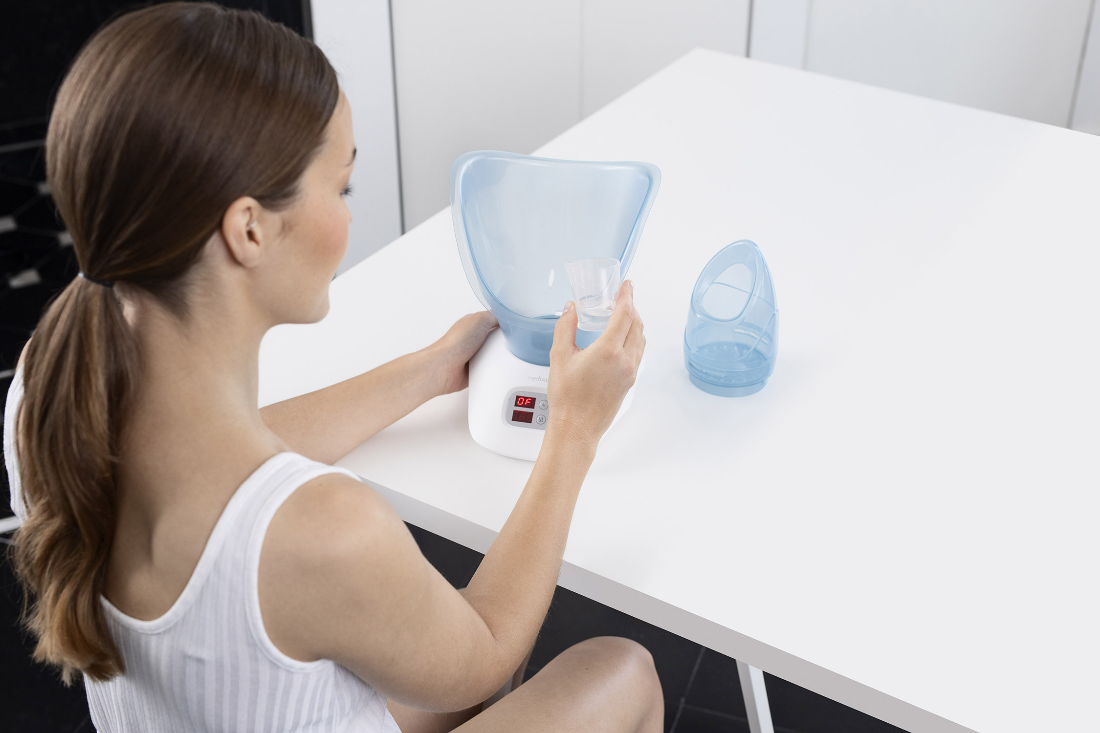 Vrouw die een gezichtsstomer gebruikt, vult de watercontainer op een witte tafel.