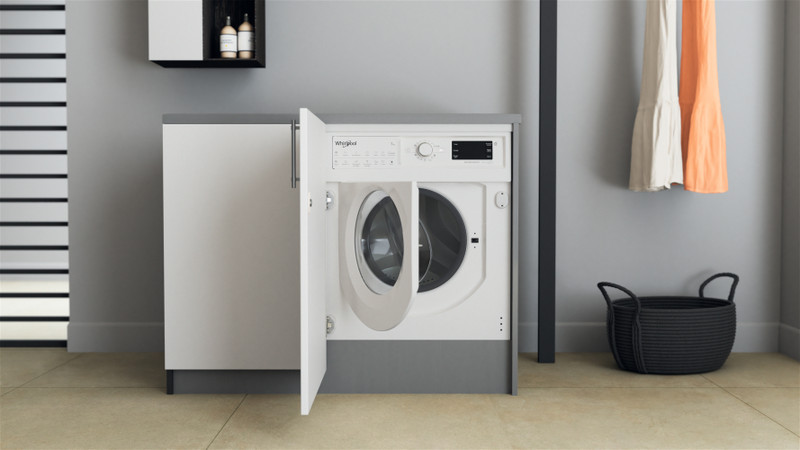 Egy fehér Whirlpool mosógép nyitott ajtóval. Balra egy szürke szekrény.