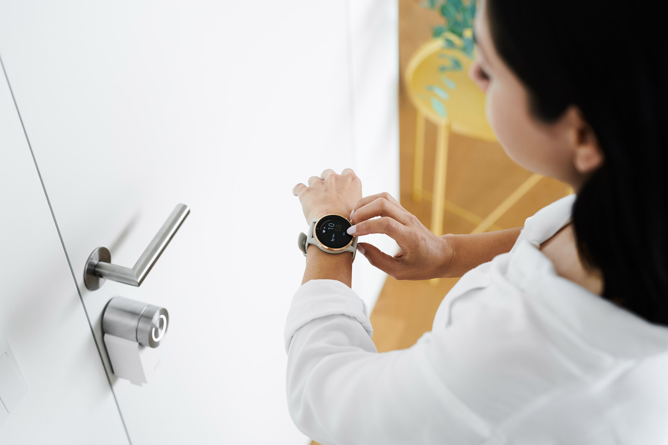 Vrouw die naar een smartwatch kijkt. Ze draagt een wit shirt. Deur en handvat zijn zichtbaar.