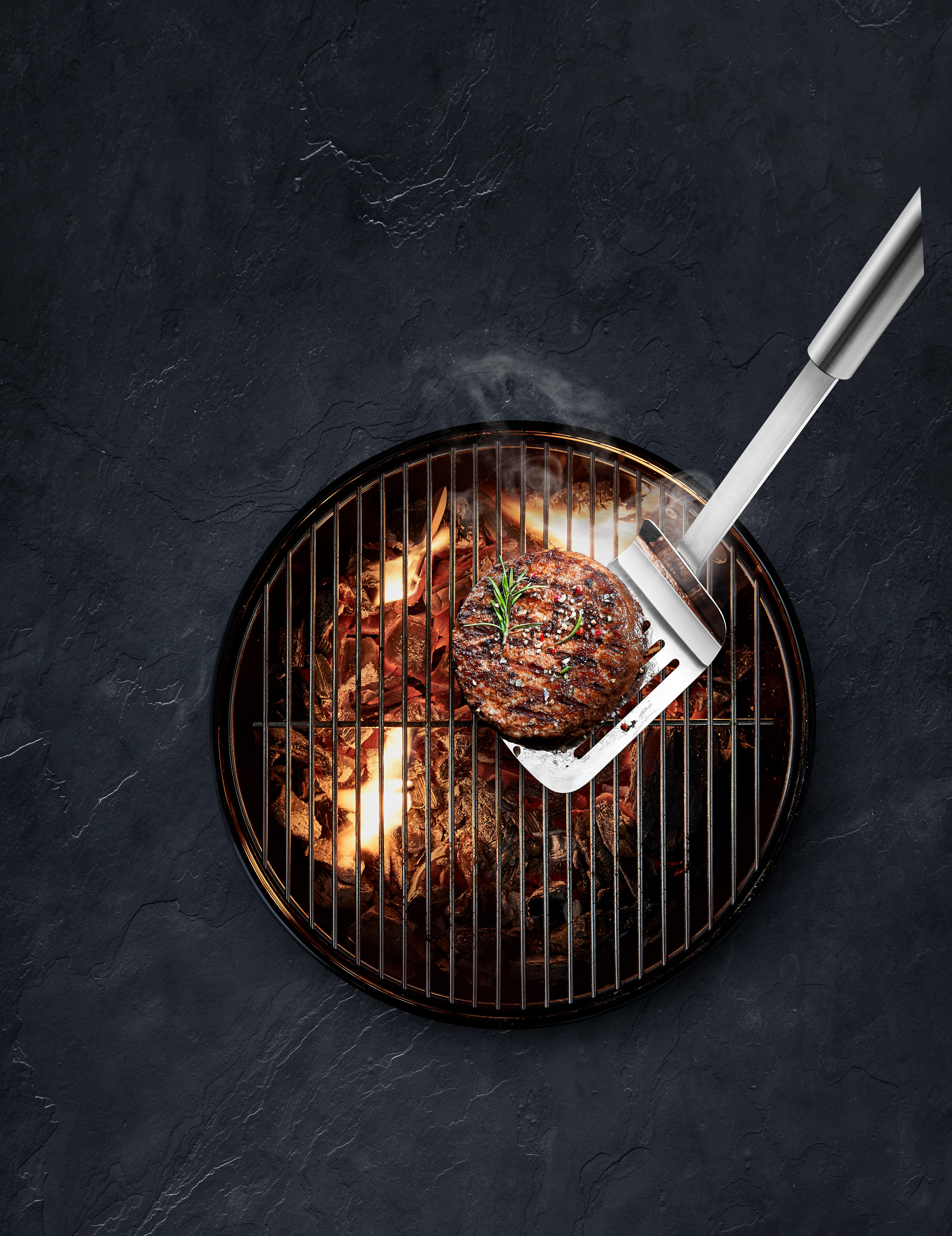 Draufsicht auf einen Burger auf einem Grill mit einem Bratenwender, auf schwarzem Untergrund.