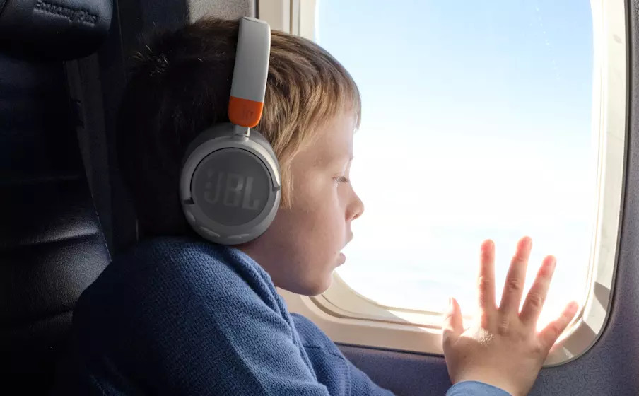 Een kind met een grijze koptelefoon kijkt uit het vliegtuigraam naar de blauwe lucht.
