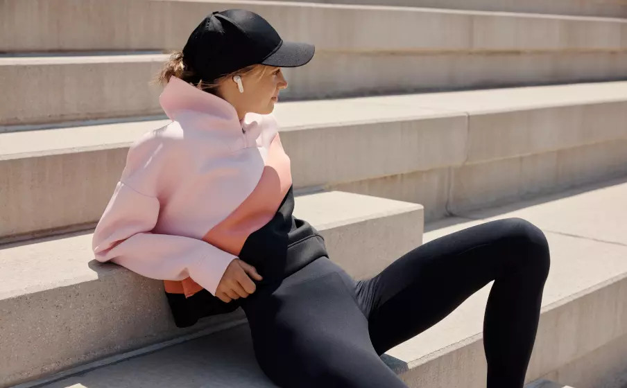 Vrouw in roze en zwarte hoodie, pet, zittend op betonnen trappen, met oordopjes.