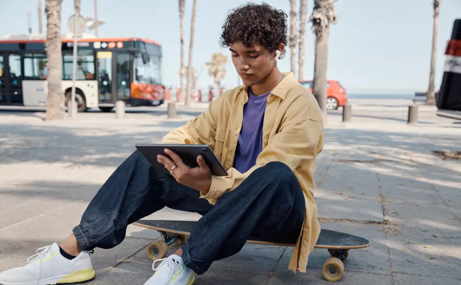 Een jonge man zit op een skateboard en gebruikt een tablet in de buitenlucht, met een bus op de achtergrond.