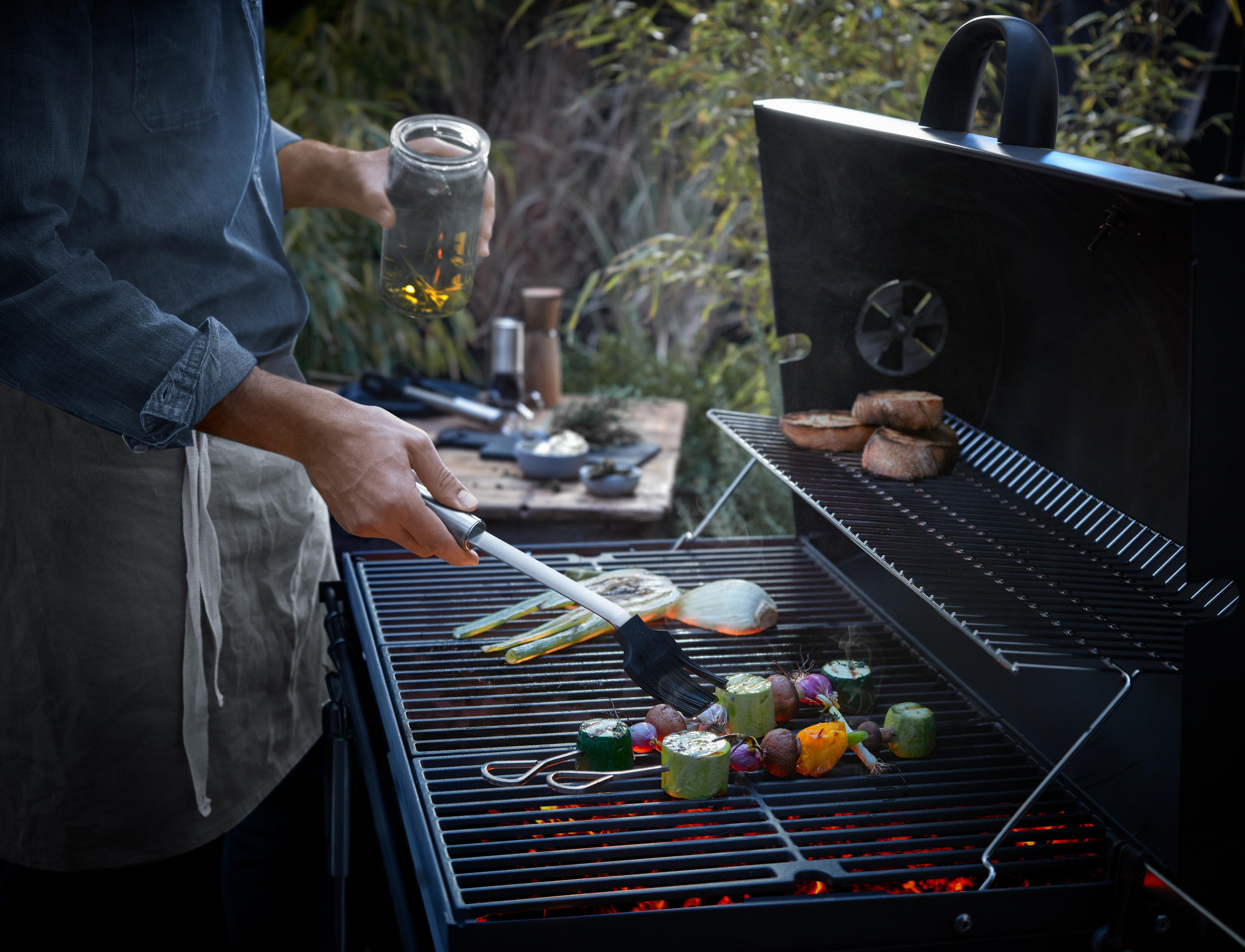 Mann grillt Gemüse auf einem Grill. Er hält einen Pinsel und ein Glas. Brot auf dem Rost.