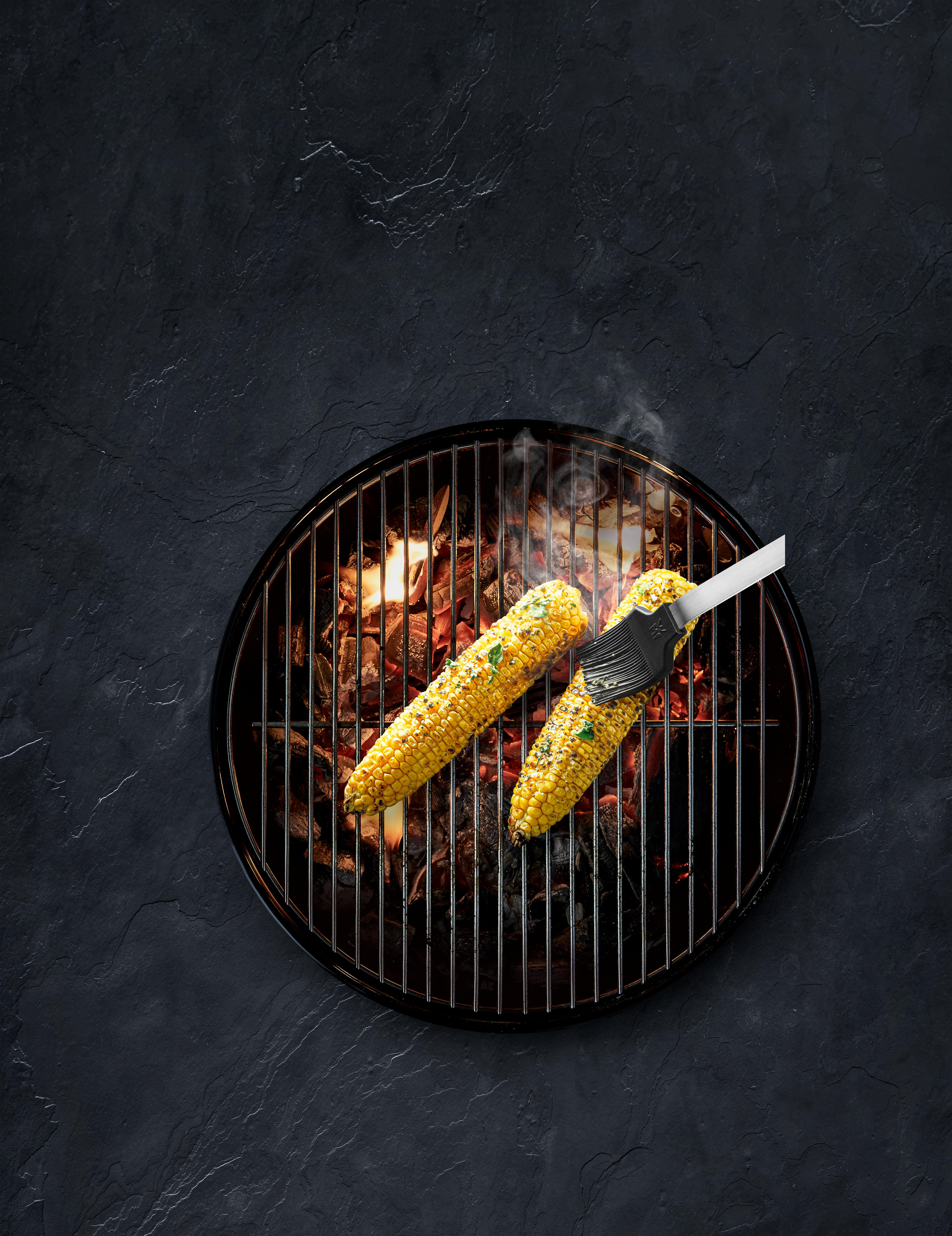 Gegrillter Maiskolben. Schwarzer Grill, dunkler Hintergrund. Butter auftragen.